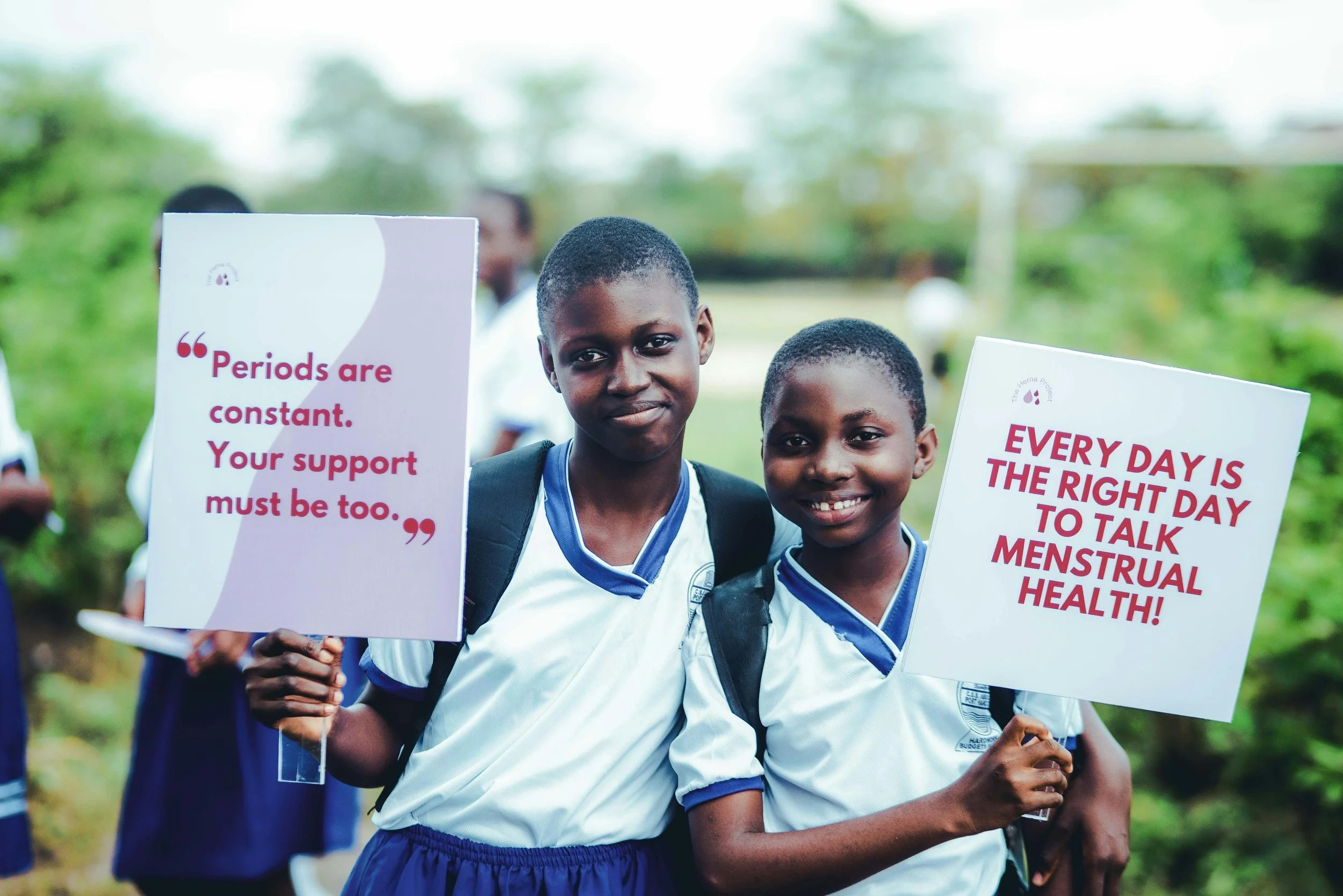 Two young girls in school uniforms holding signs about menstrual health awareness during an outdoor event.