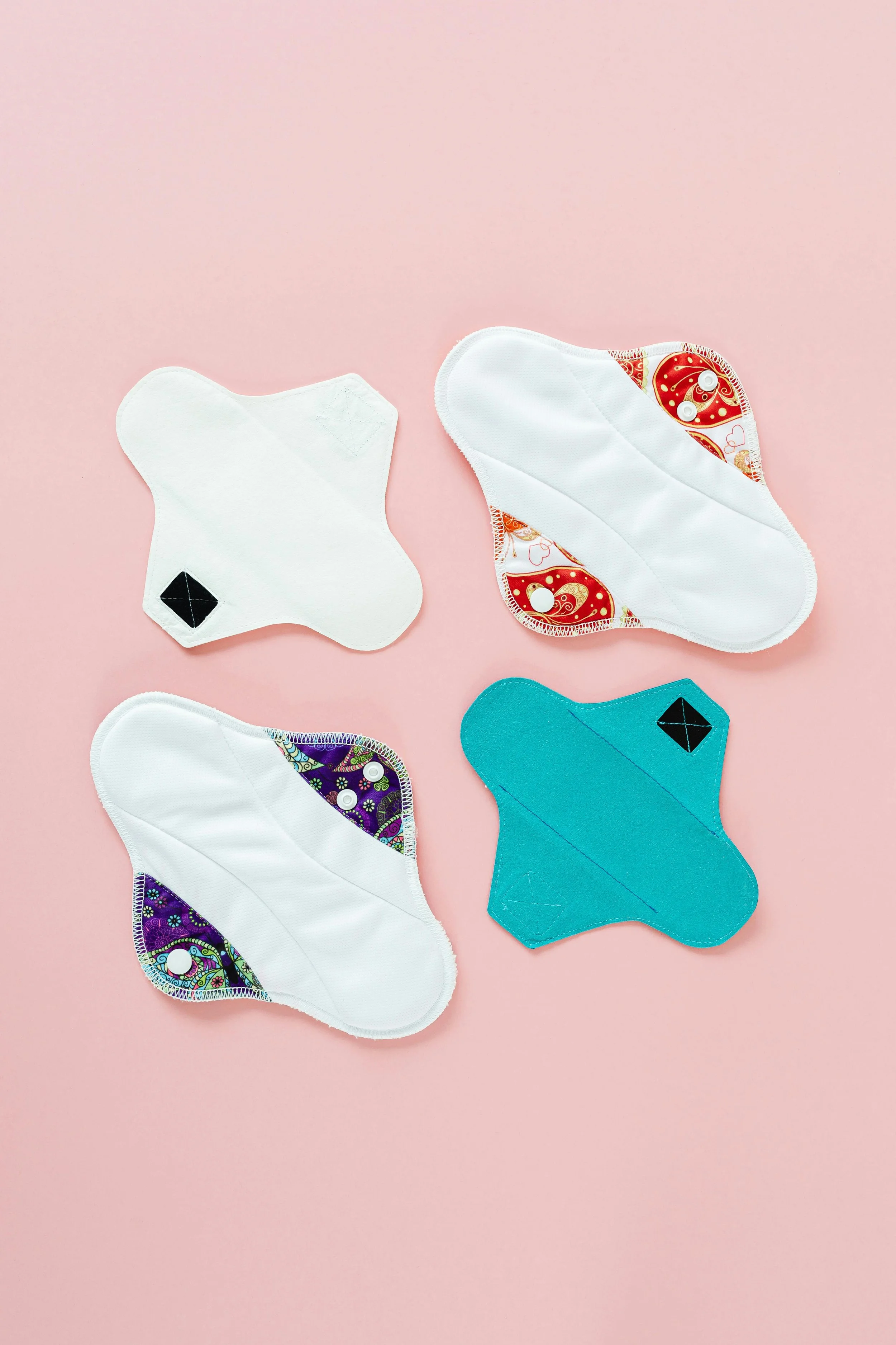 Several reusable cloth menstrual pads with colorful patterns and a solid teal pad on pink background.