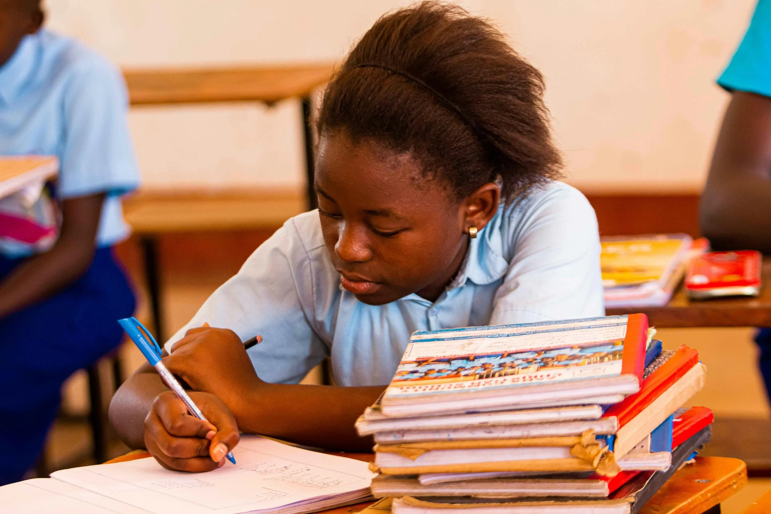 A girl with a high puff hairstyle is writing in a notebook at a desk filled with colorful textbooks in a classroom.