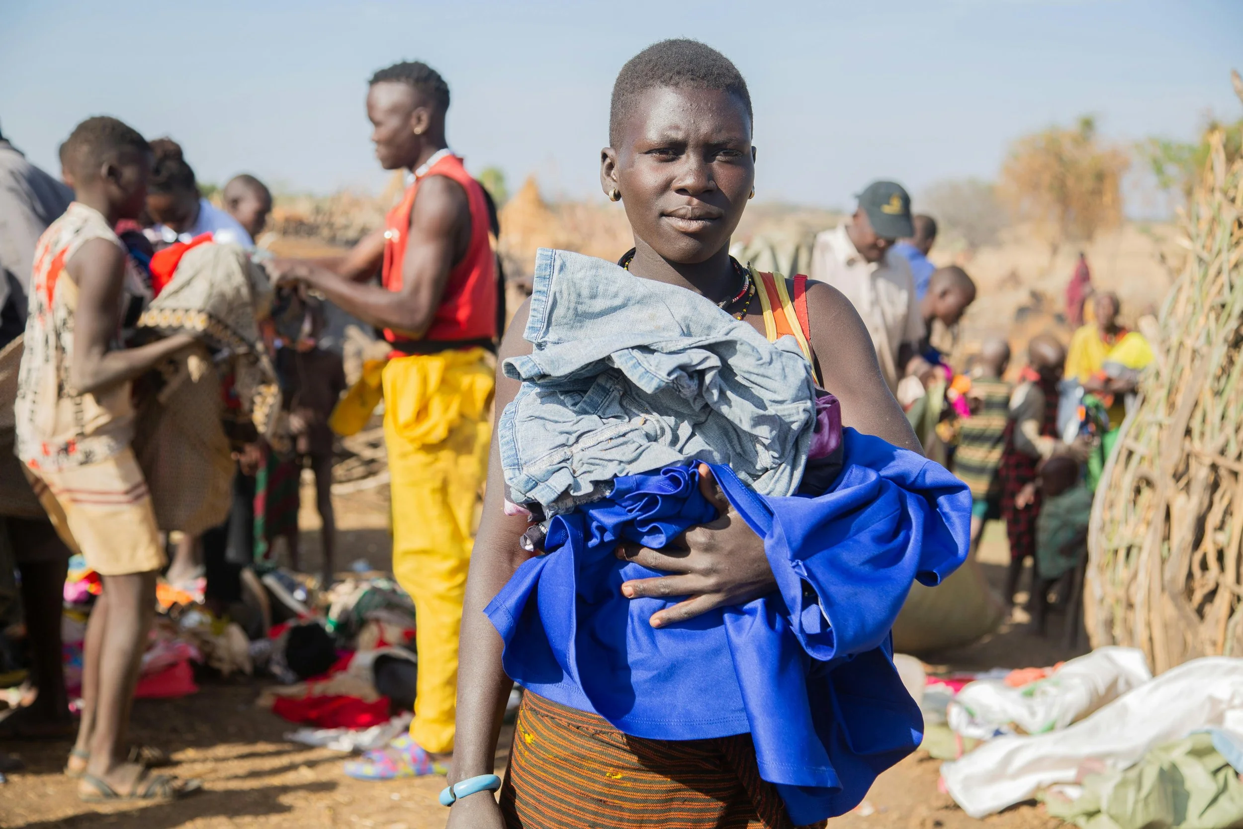 A young woman with short hair and dark skin holding clothing in front of a market scene with other people and stalls in the background.