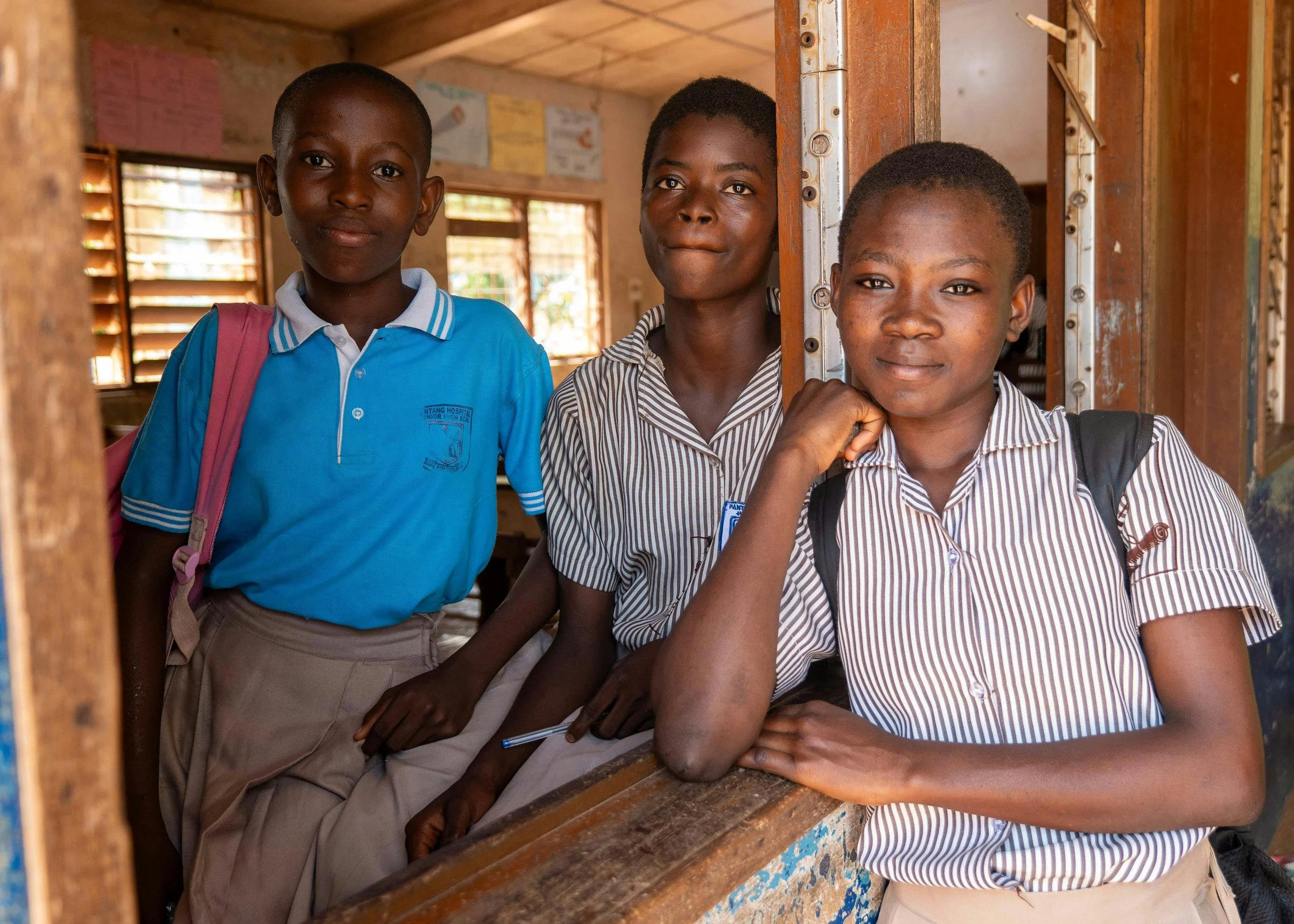 Three children wearing school uniforms, standing inside a classroom and looking at the camera.