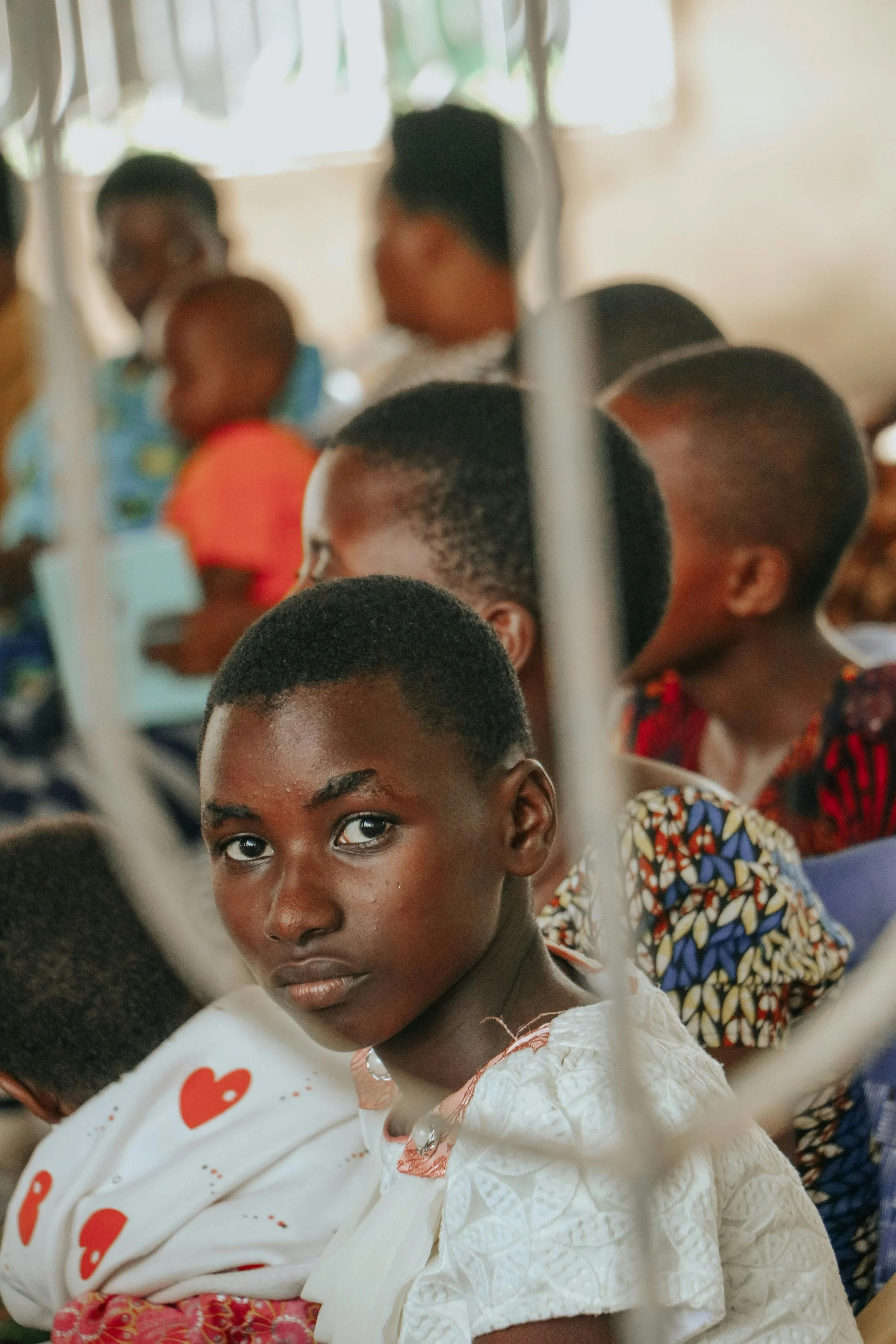 A young girl with dark skin and short hair looking into the camera, seated indoors with a group of people in the background.