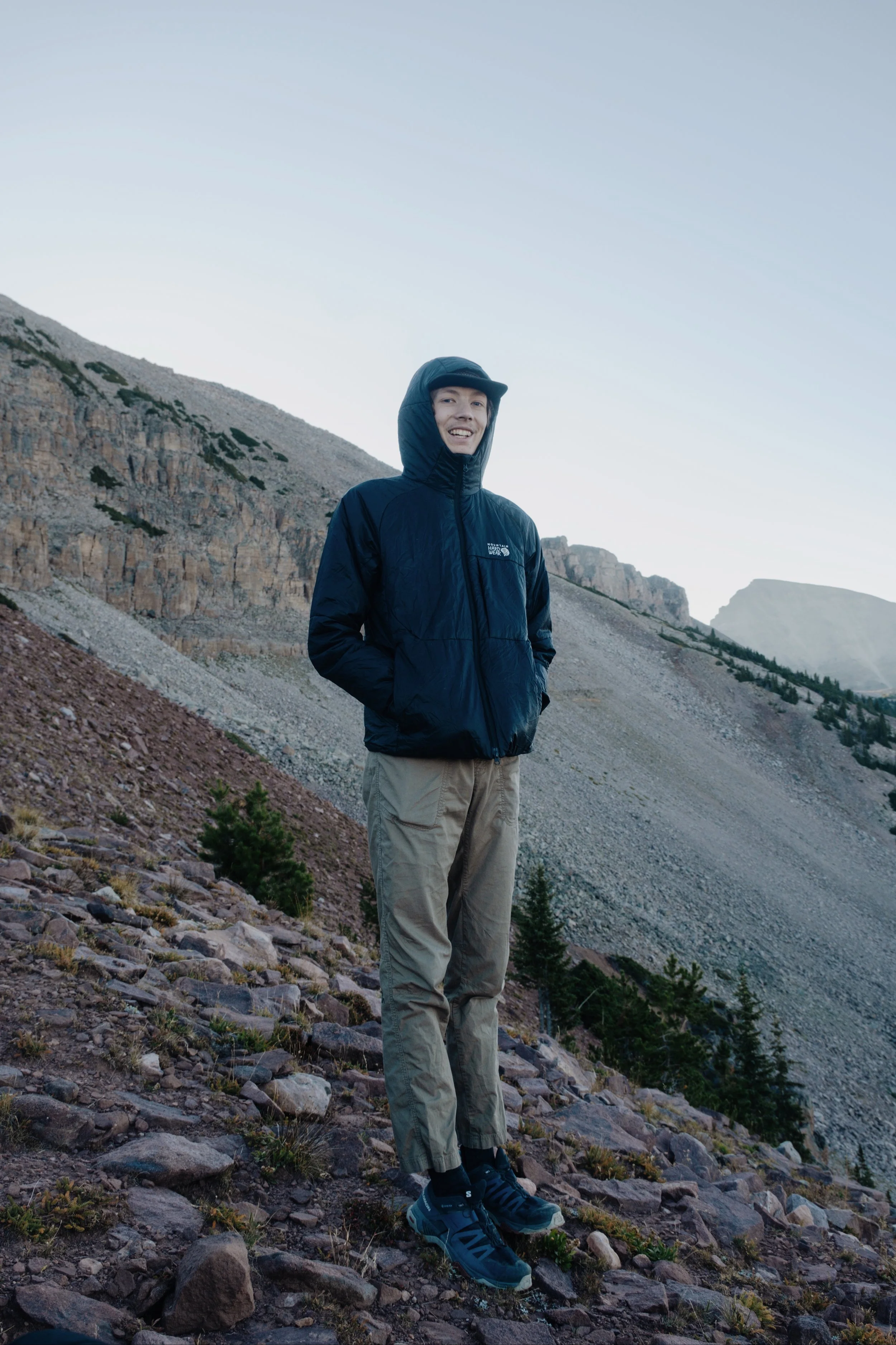 A young man wearing a black hooded jacket and beige cargo pants standing on a rocky mountainside with a clear sky in the background.
