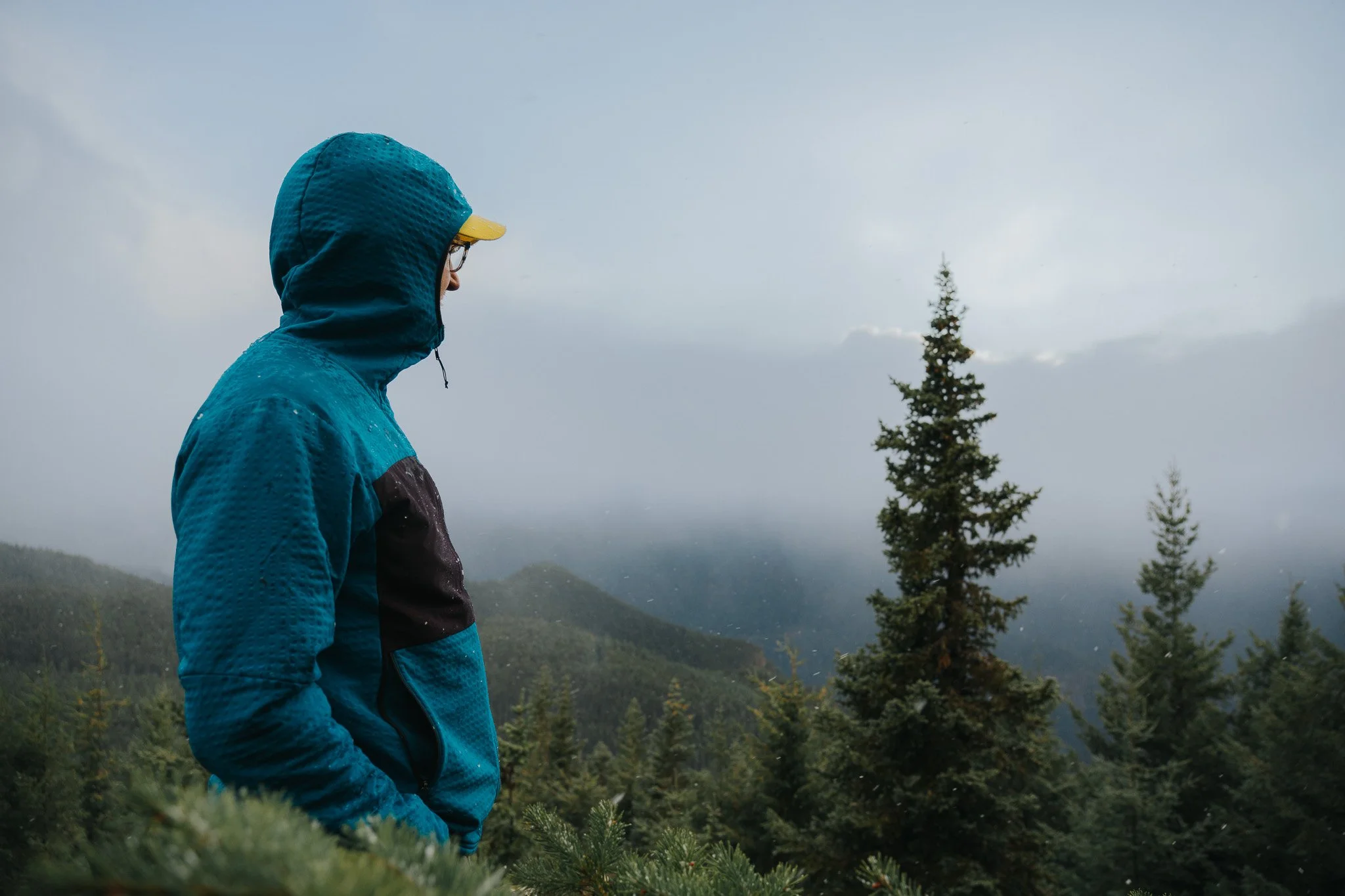 A person dressed in a blue rain jacket with the hood up, wearing glasses and a yellow cap, standing in a misty, forested mountainous area during rainy weather.