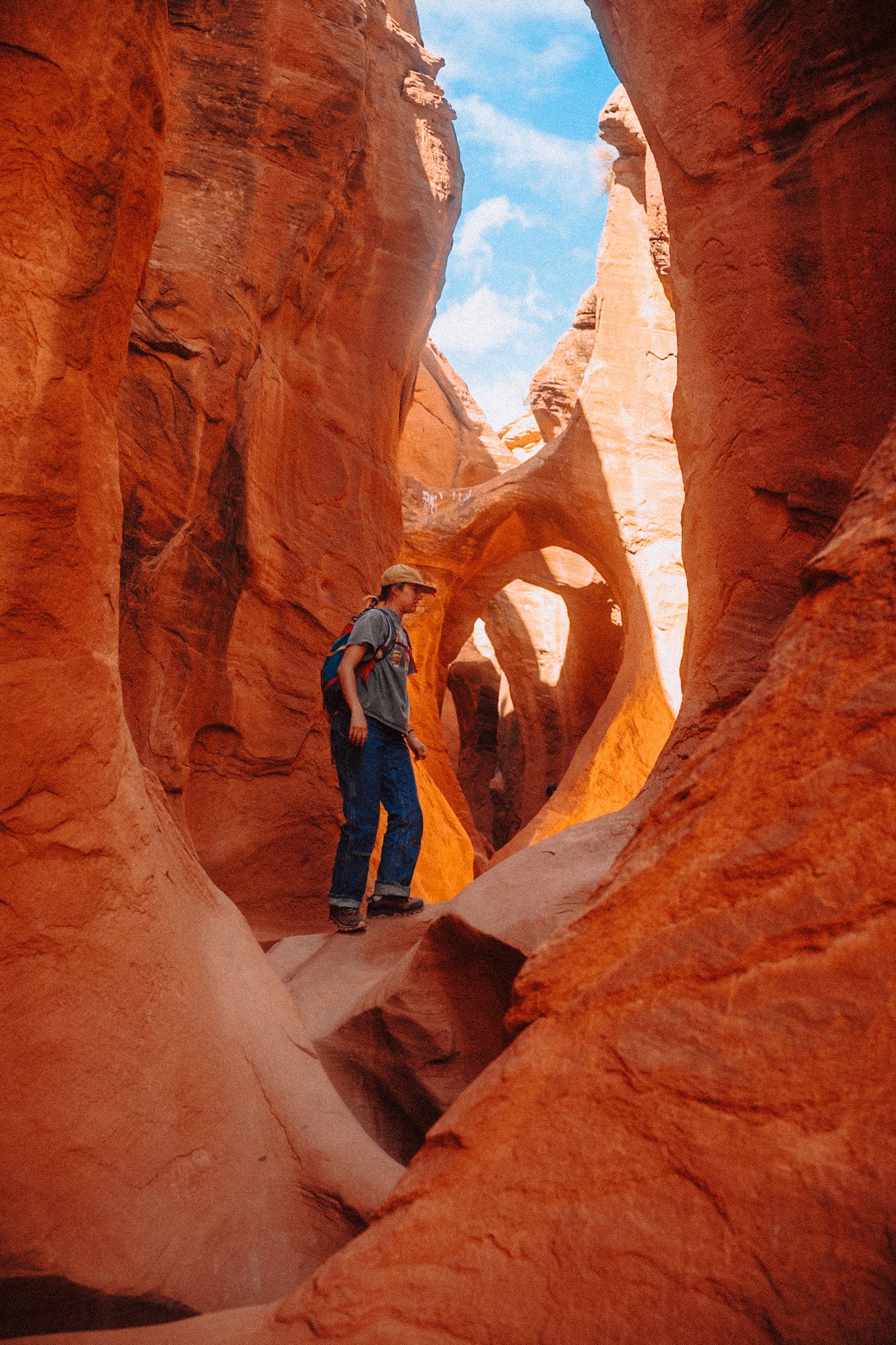 A person hiking through a slot canyon with orange rock walls and a blue sky with clouds overhead.