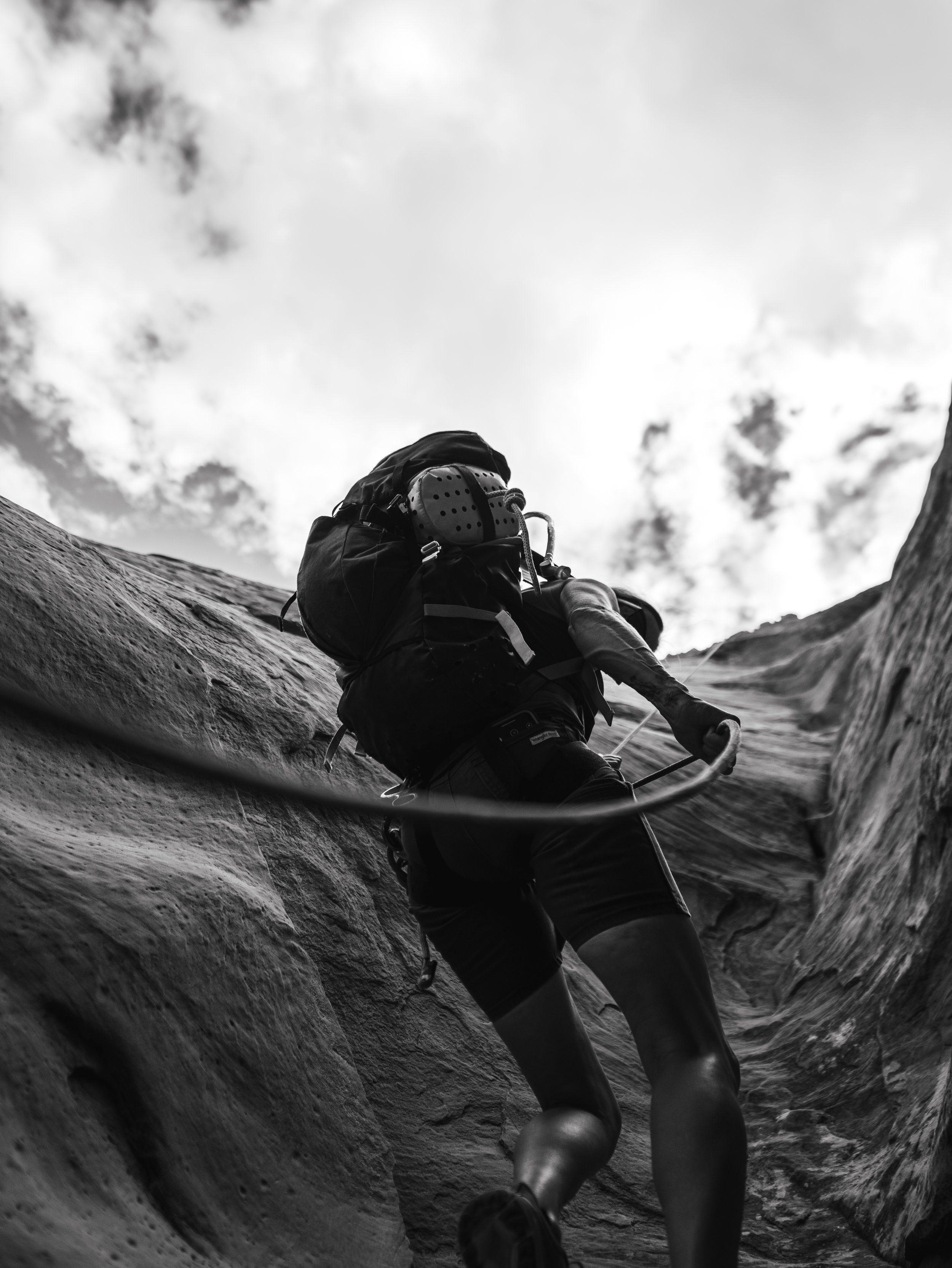 A person climbing a rocky canyon wall, equipped with a backpack and climbing gear, black and white photo