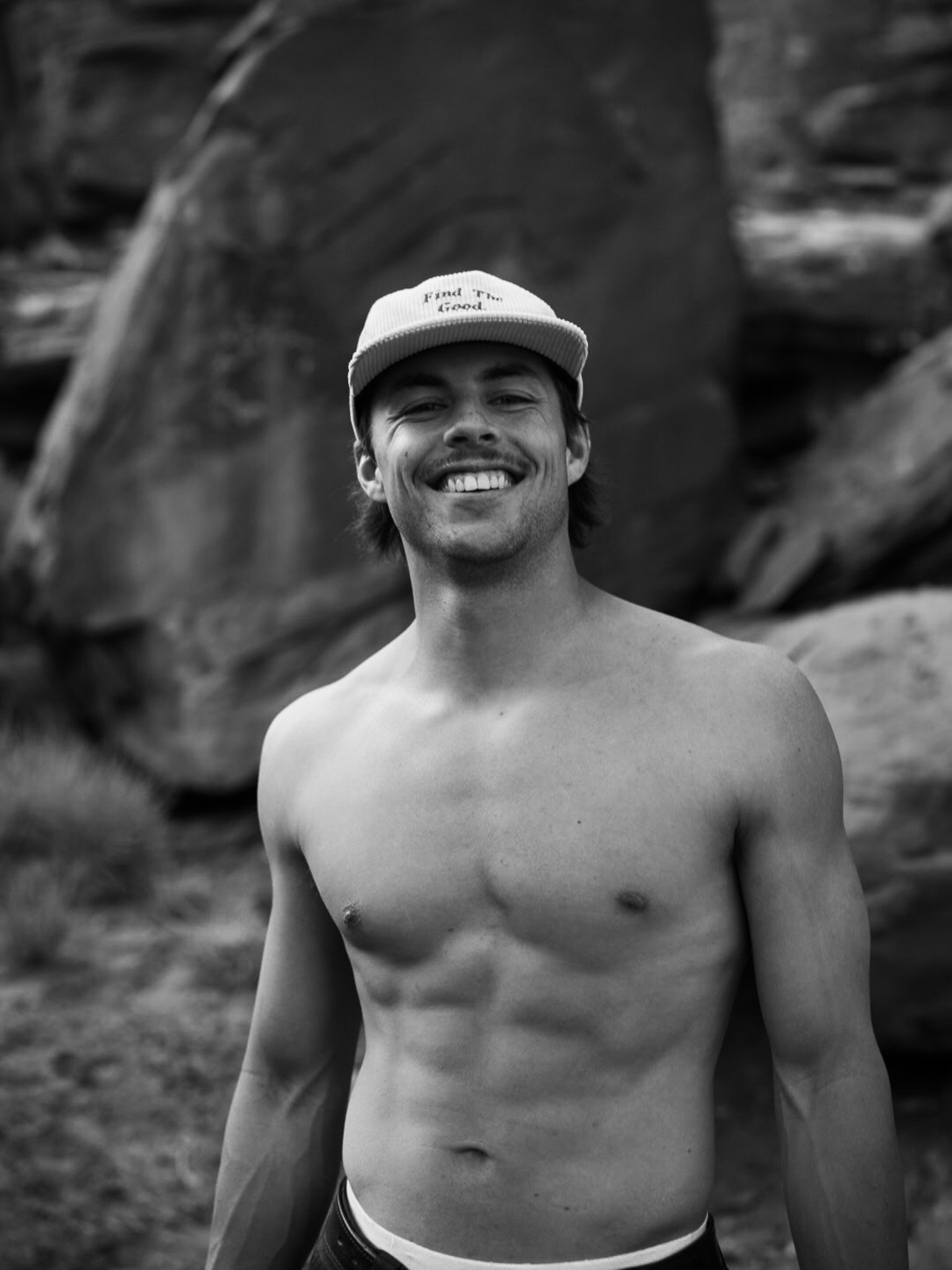 Black and white photo of a shirtless young man smiling at the camera, wearing a baseball cap, outdoors with rocks in the background.