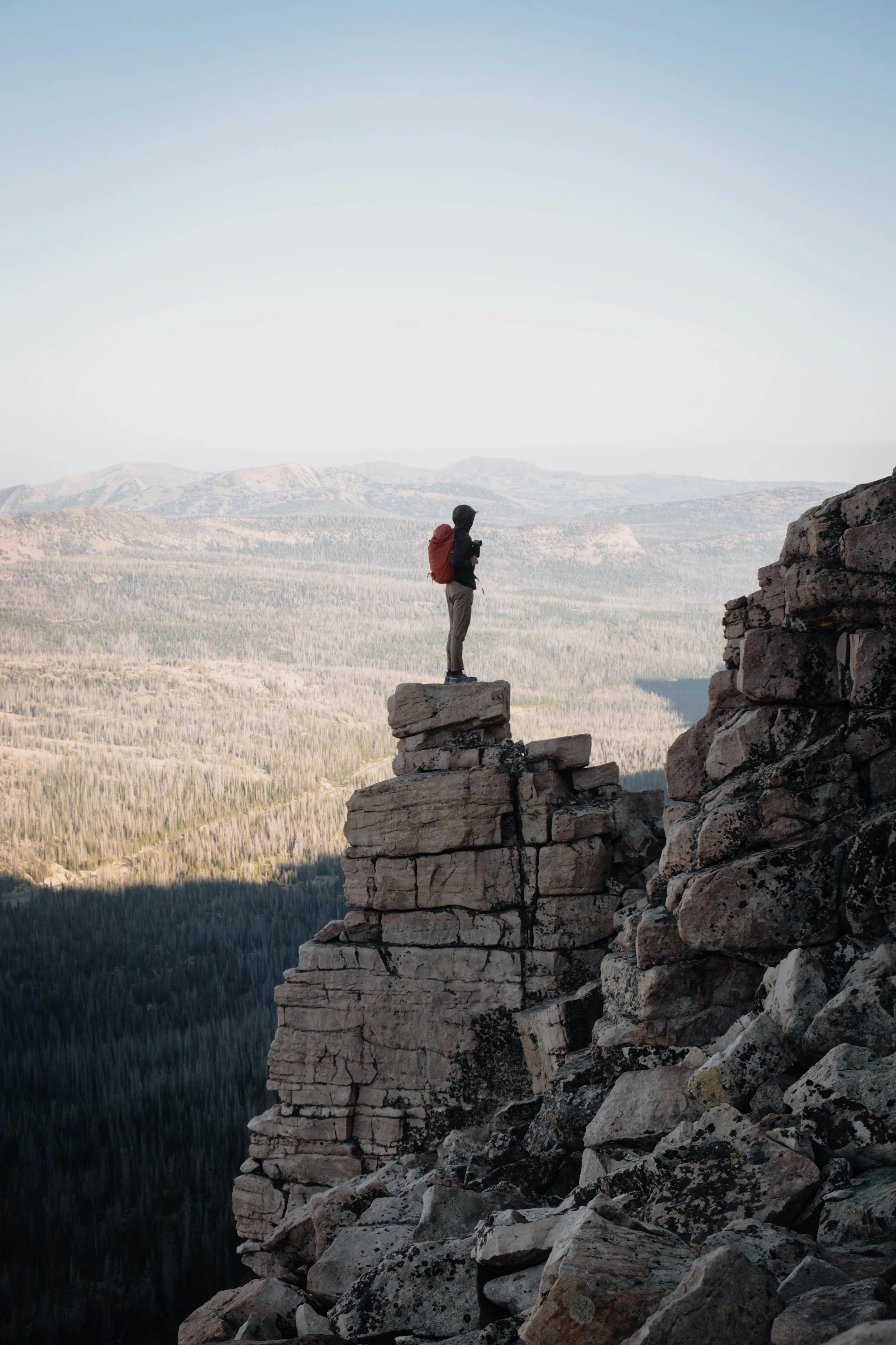 Person standing on a large rock ledge overlooking a vast, mountainous landscape during daytime.