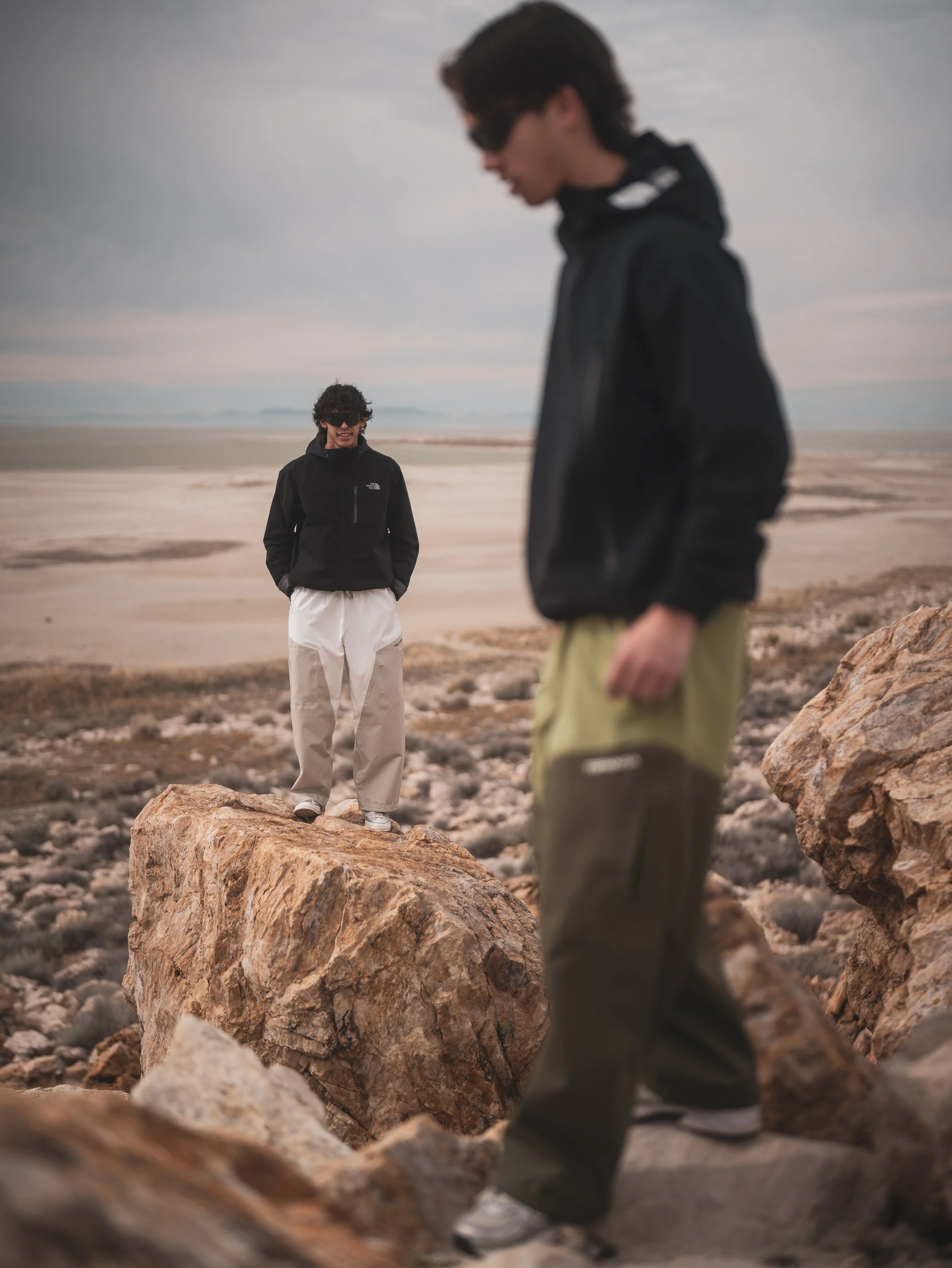 Two young men outdoors on a rocky terrain, one in focus standing on a large rock, smiling and looking at the other, who is in the foreground with his head down and blurred. The sky is overcast, and distant mountains can be seen in the background.