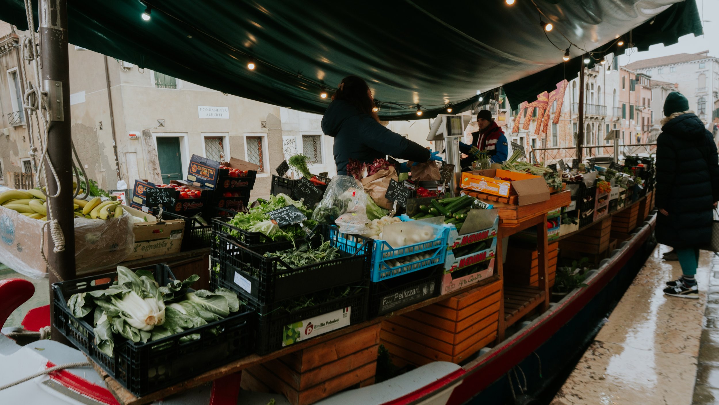 Outdoor market stall with fresh vegetables and fruits, with vendors and customers, under a green canopy, on a rainy day in an old European city.