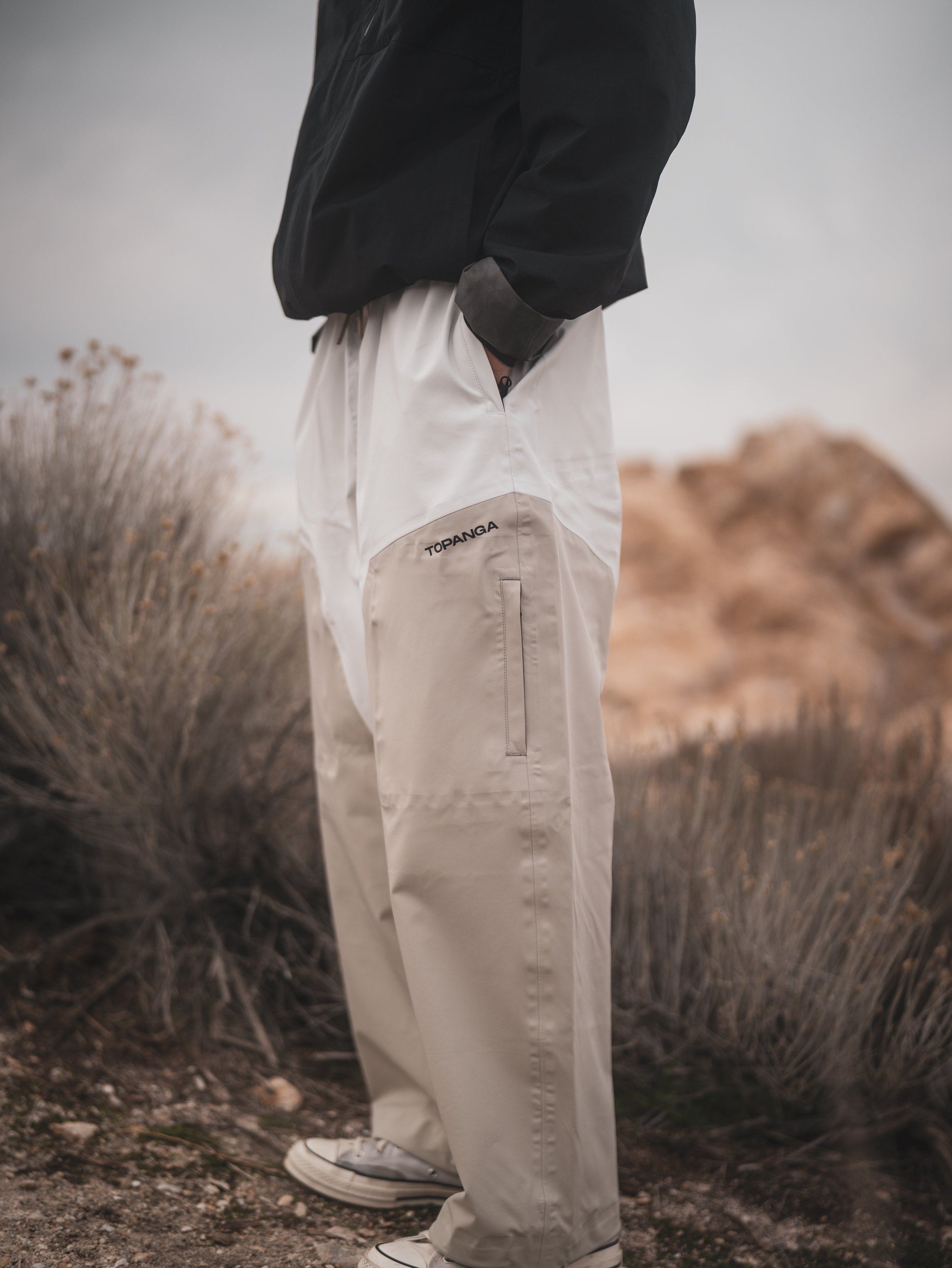 Close-up of a person wearing beige cargo pants with the brand name 'TOPANGA' on the pocket, standing outdoors in a rocky, desert-like environment.