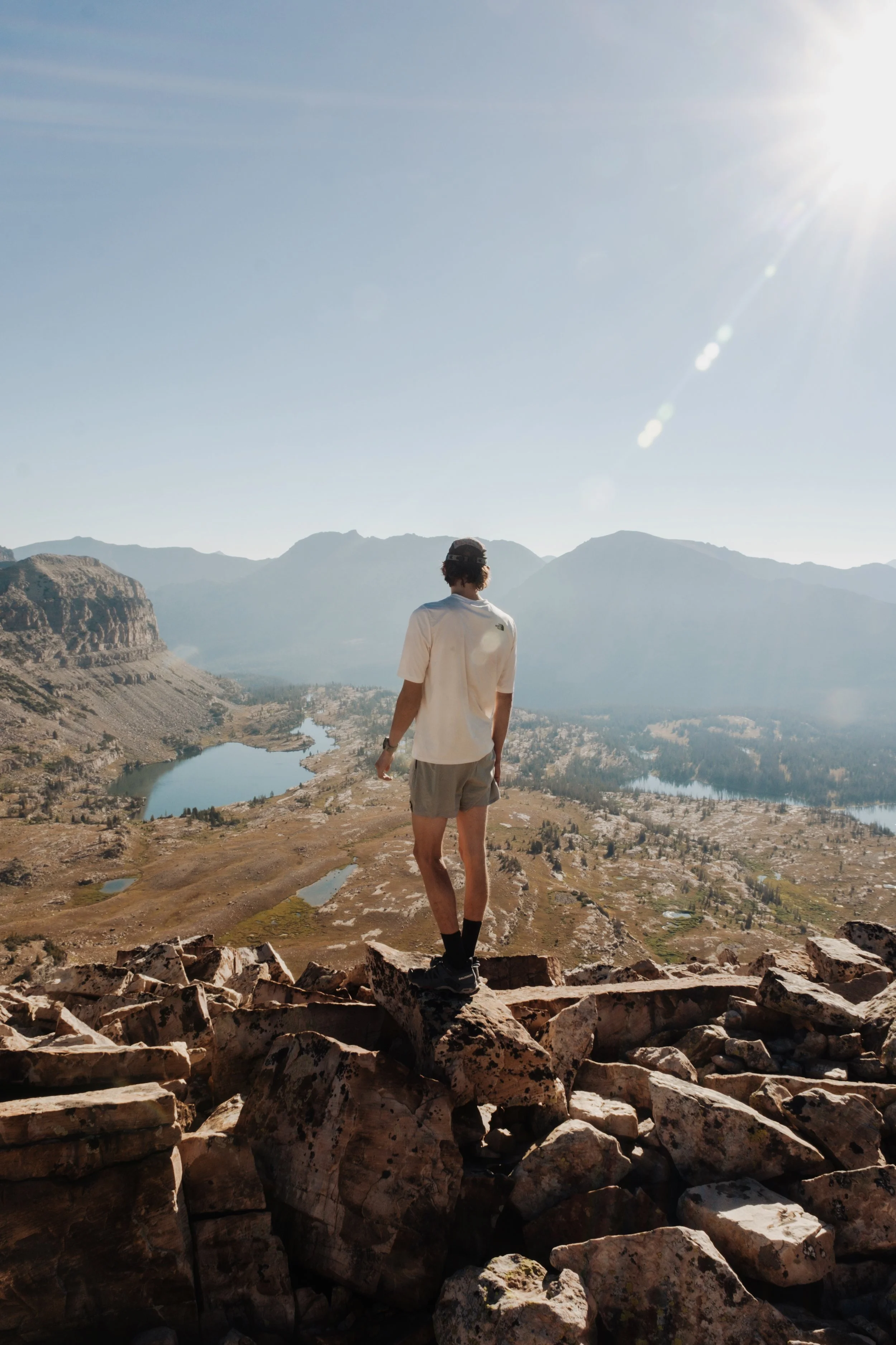 Man standing on rocks overlooking a valley with lakes, mountains in the background, and the sun shining brightly in the sky.