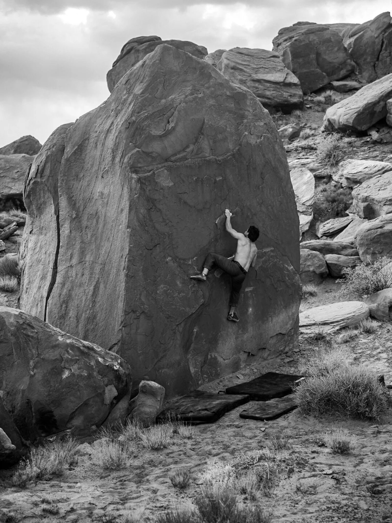 A shirtless man wearing pants and climbing shoes bouldering on a large rock in a desert area with scattered bushes, surrounded by other rocks and boulders.