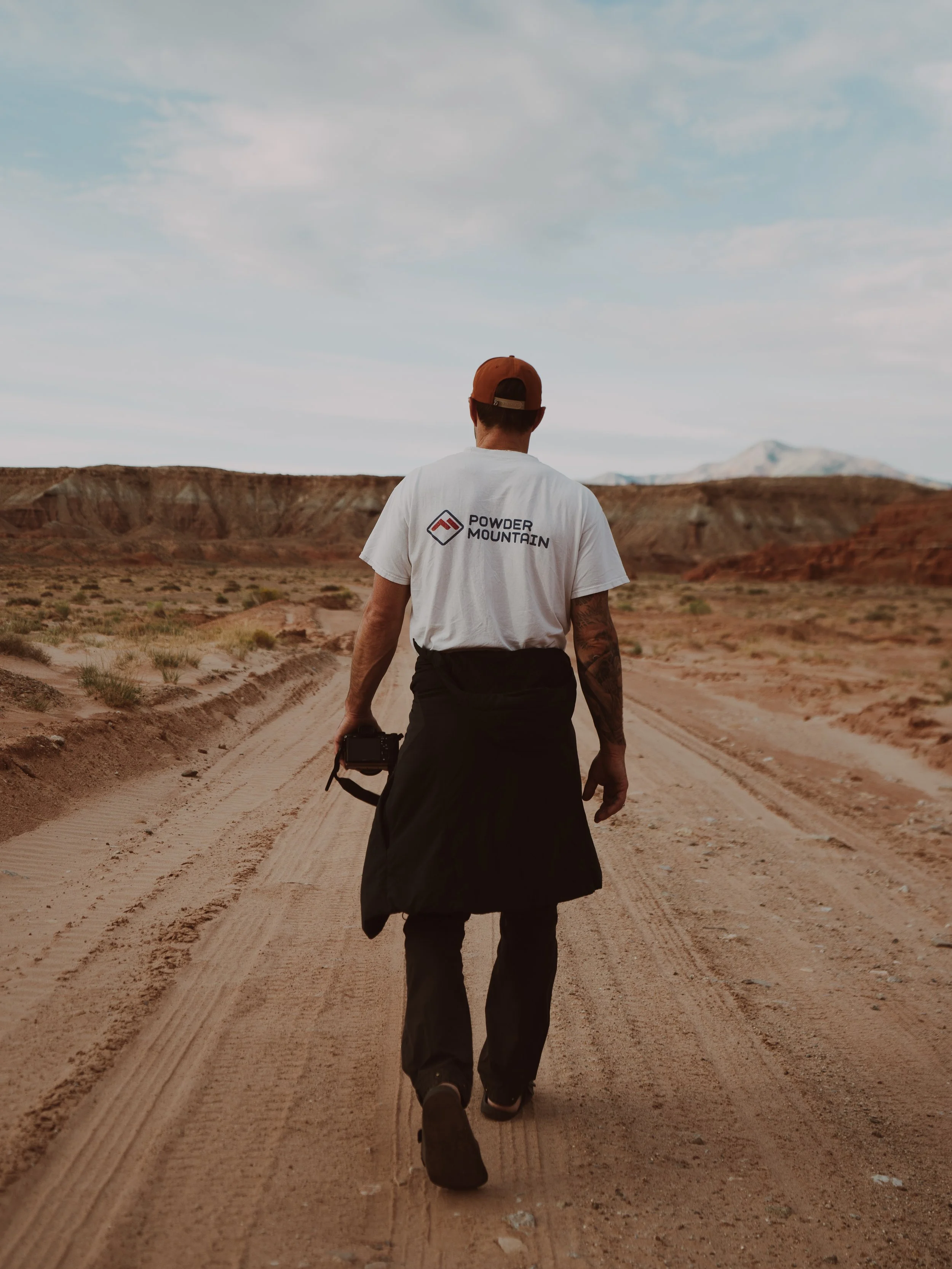 Person walking on dirt road in desert landscape, wearing a white t-shirt, black pants, brown cap, and carrying a camera, with mountains in the background.