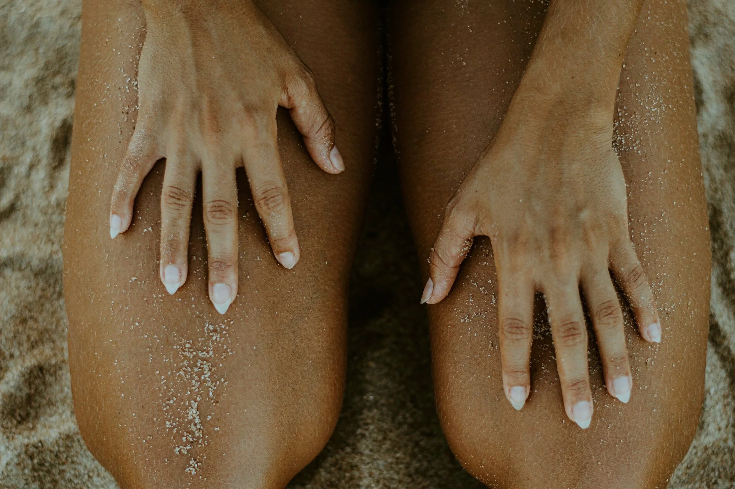 Close-up of a person's spray-tanned hands resting on their thighs on a sandy surface, with sand on their skin and fingers.
