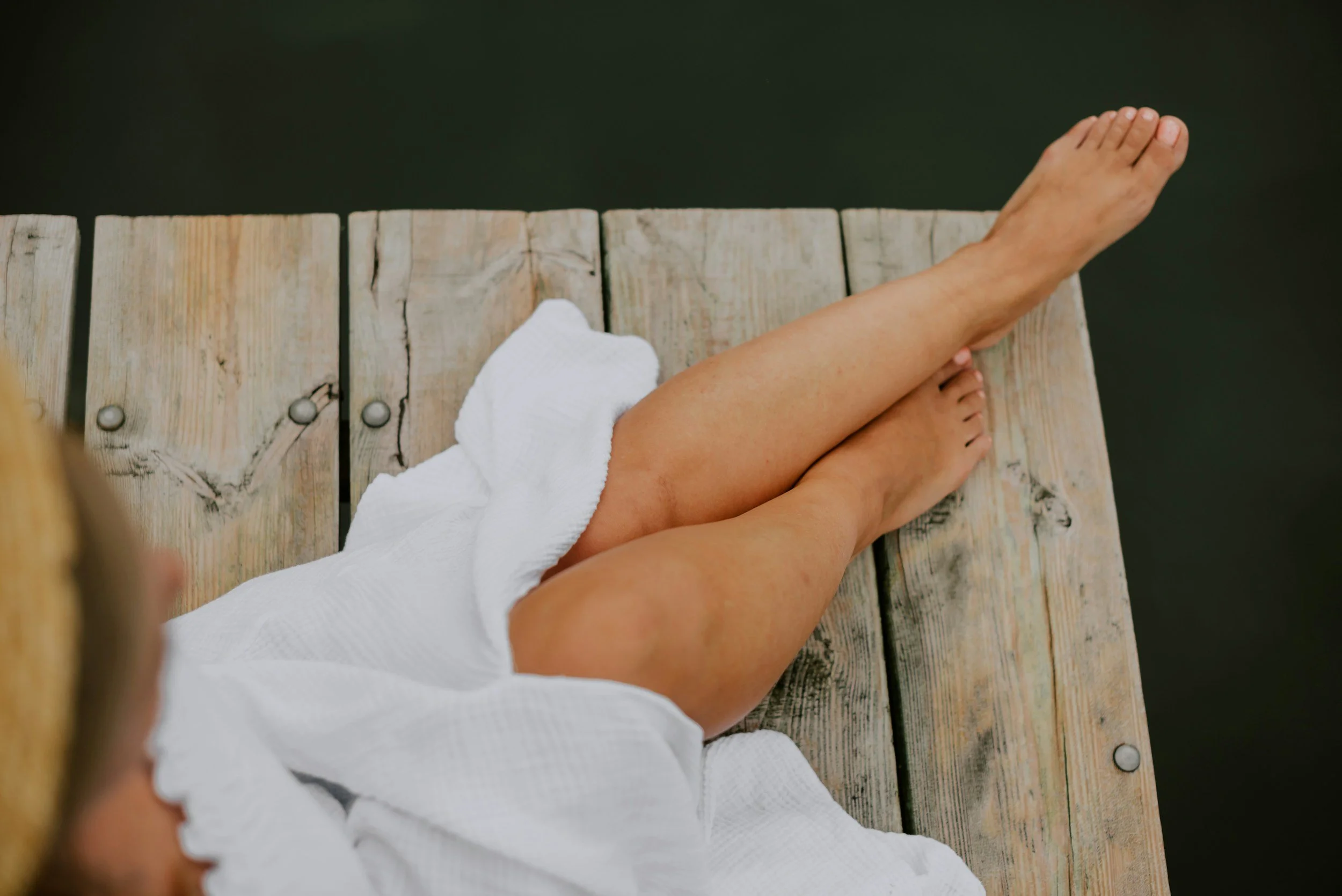 Person with tanned legs wearing a white towel sitting on a wooden dock over dark water.