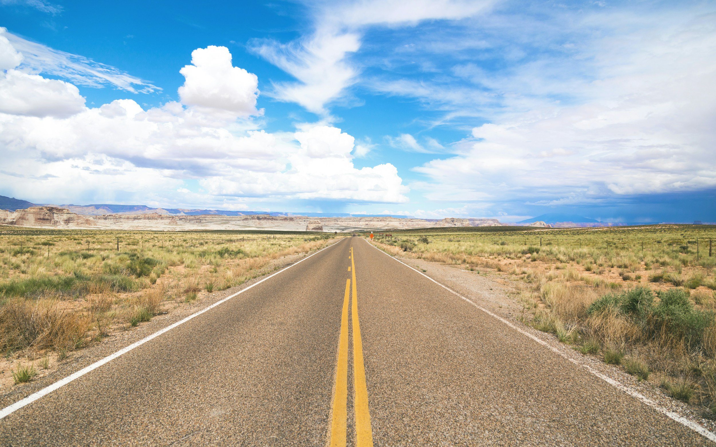 Long straight road with yellow dividing lines in a desert landscape, with distant mountains and a partly cloudy sky.