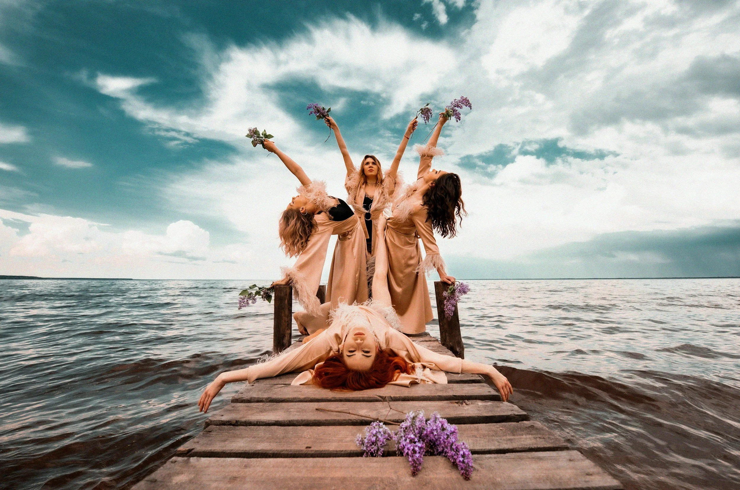 Five women are posing on a wooden dock by the water, with a cloudy sky in the background. They are dressed in beige and black clothing with feathered sleeves, holding lavender flowers, some with arms raised, some lying down, creating a artistic arrangement.