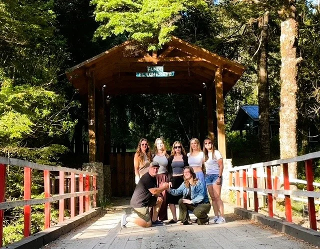 Group of six women and two men posing on a wooden covered bridge surrounded by green foliage