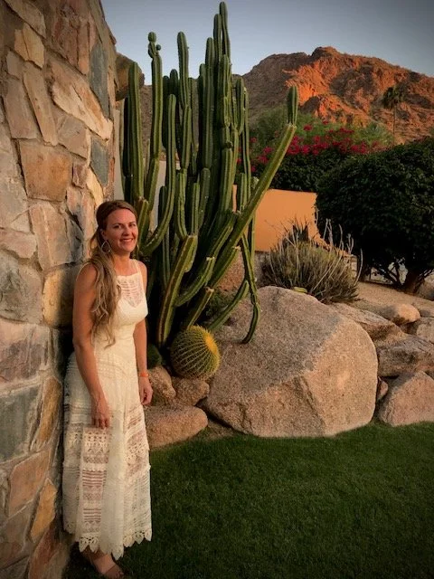 Founder of Sunflower Tans in a white dress standing next to a tall cactus outdoors, with desert landscape and mountains in the background during sunset.