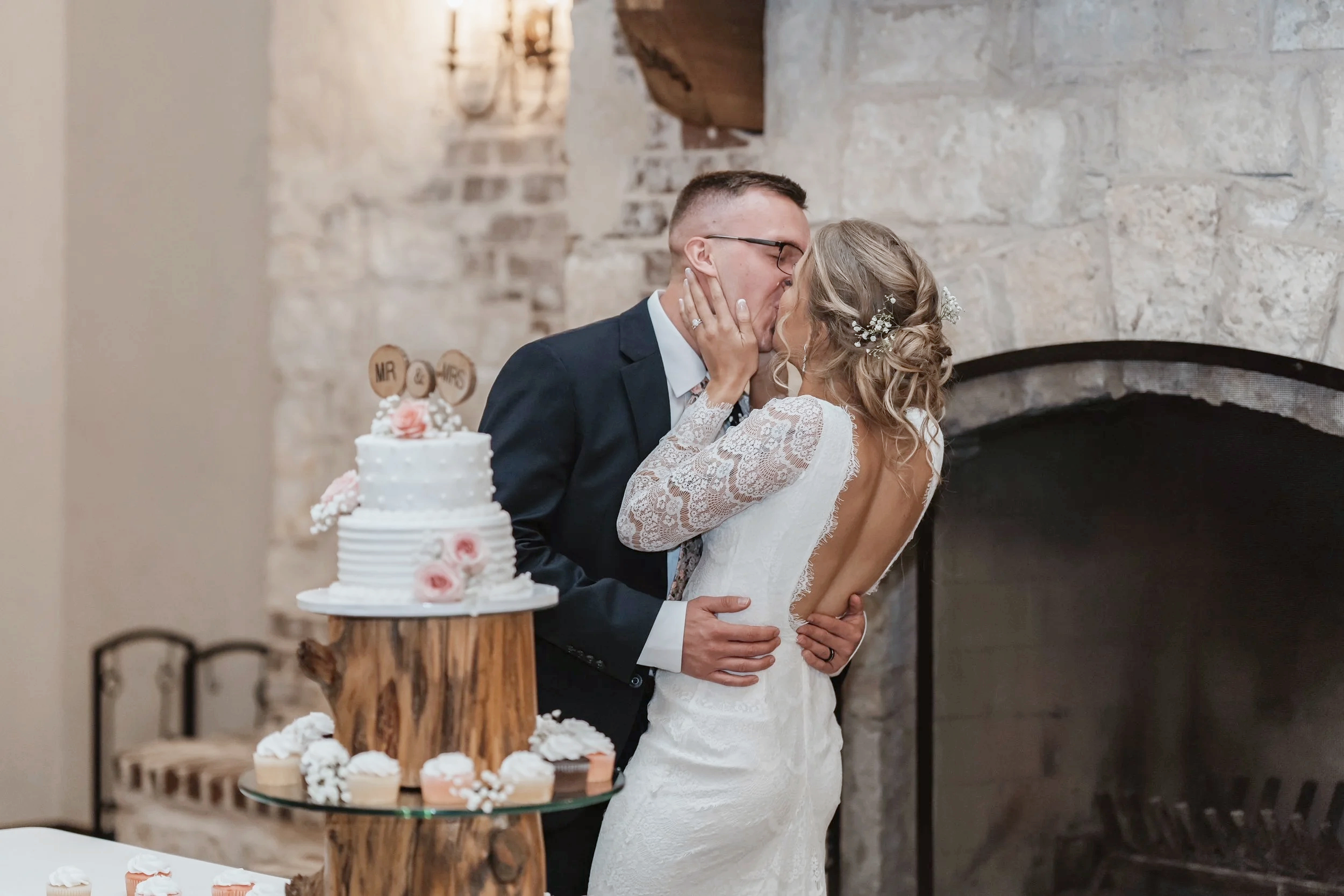 A couple sharing a kiss at their wedding reception, with a wedding cake decorated with pink flowers and a wooden table in the foreground, and a stone fireplace in the background.