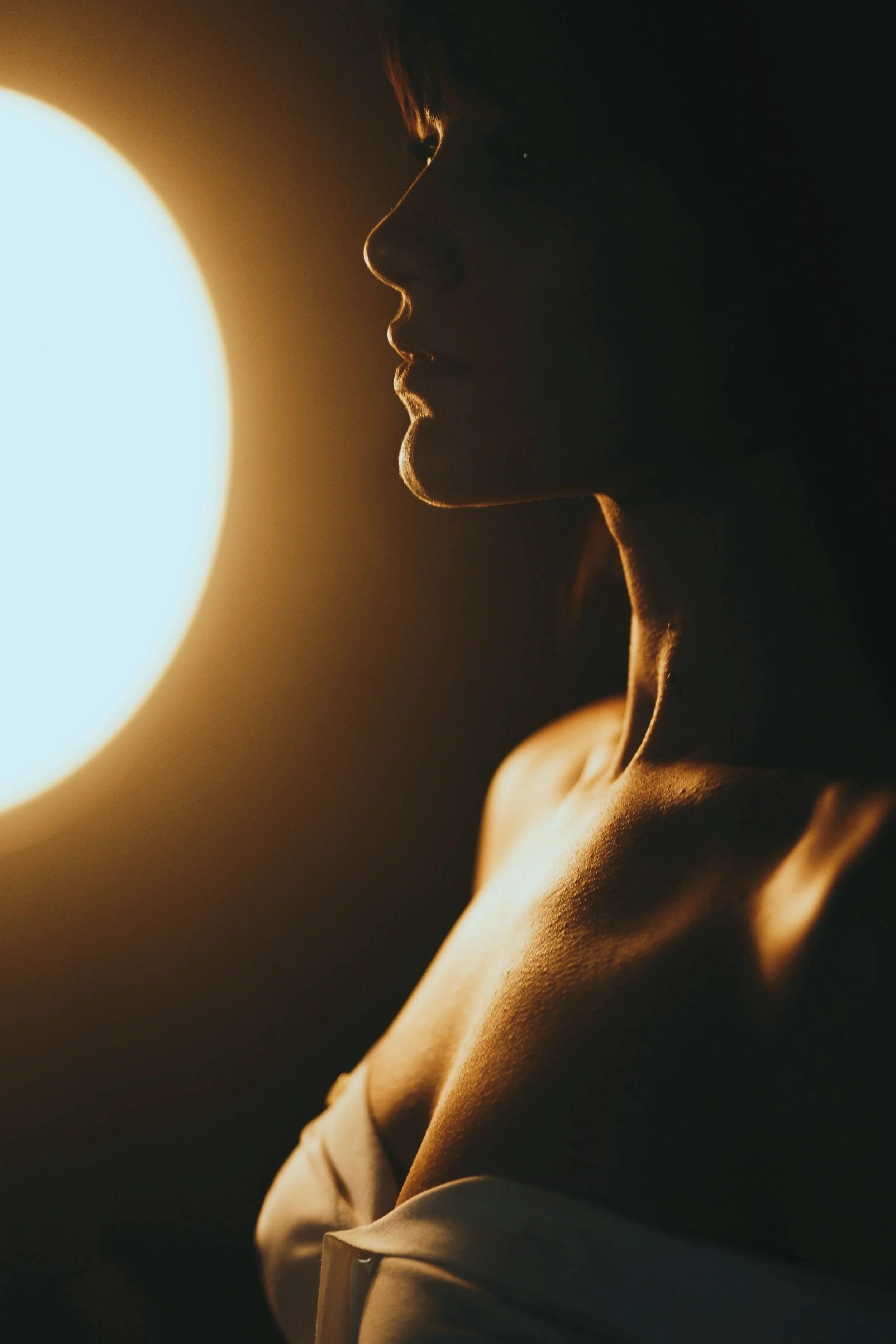 Side profile of a woman with short hair, illuminated by warm light, against a dark background.