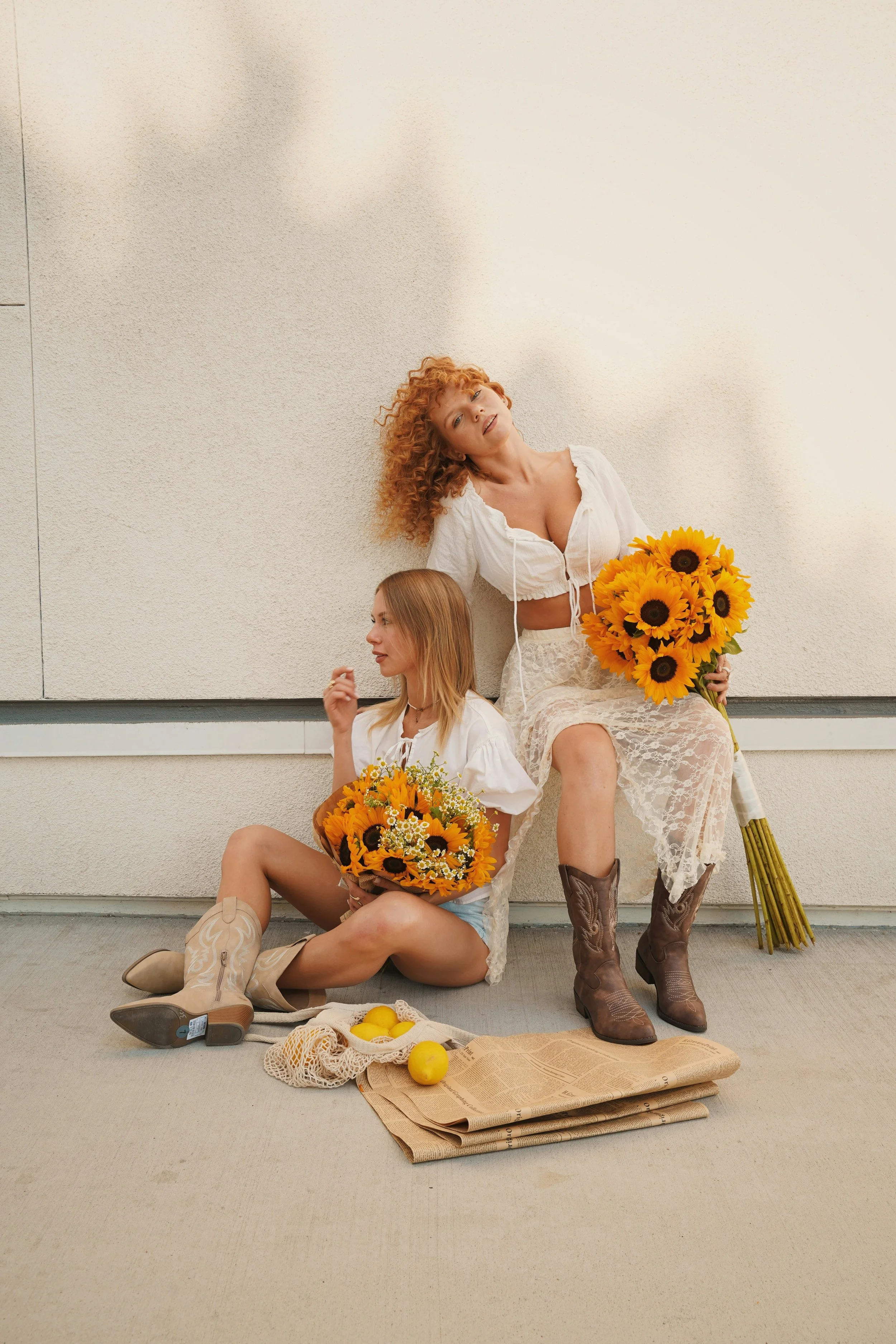 Two women sitting on the ground with sunflowers, lemons, and a paper bag, against a plain wall background.