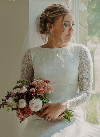 A bride holding a bouquet of pink and white flowers, standing by a window, wearing a lace wedding dress with her hair styled in an updo.