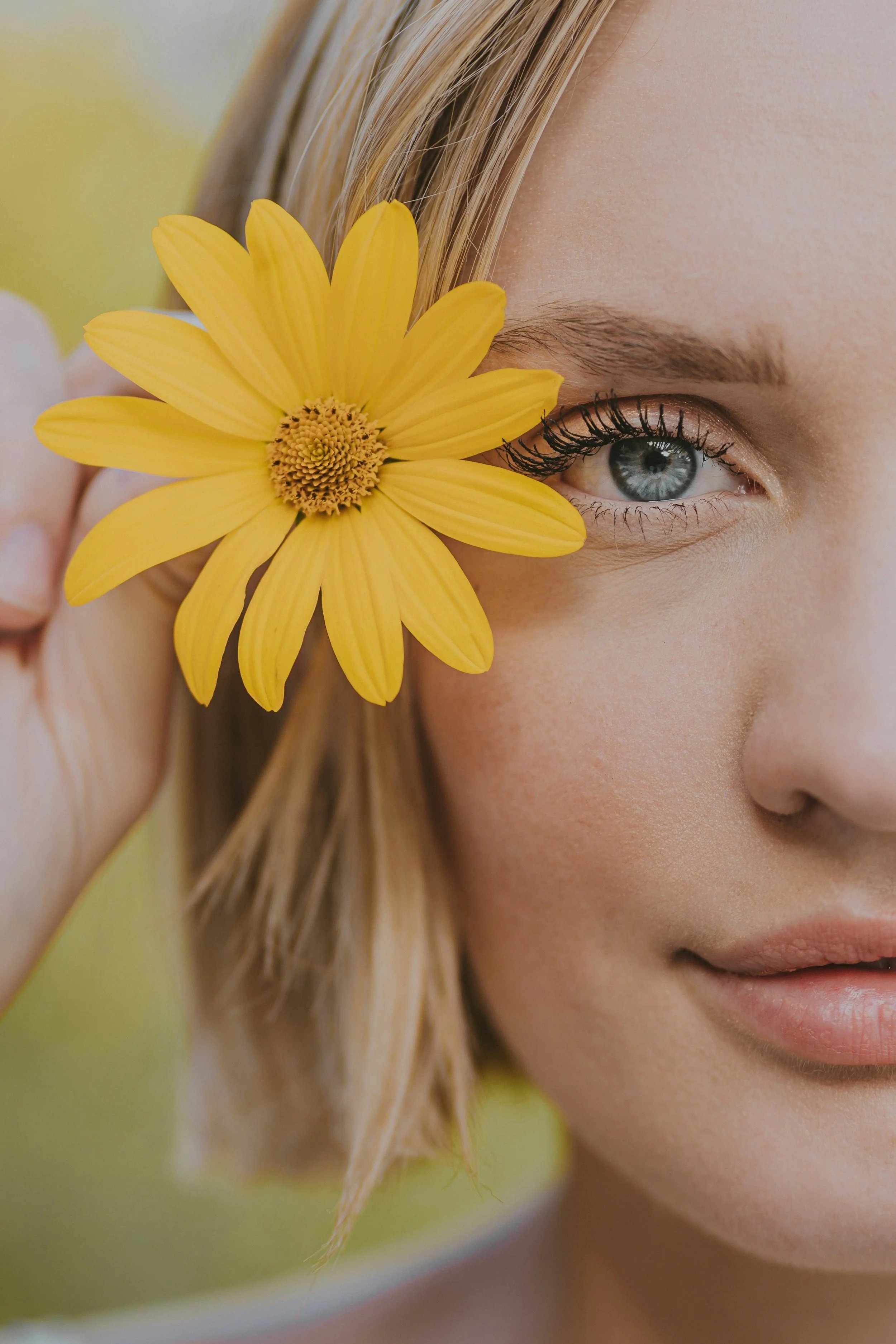 Close-up of a woman's face with a yellow flower covering part of her eye and cheek, showcasing her eye and skin texture.