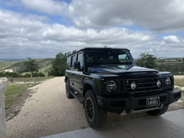 A black Land Rover Defender SUV parked on a gravel driveway with a scenic landscape of trees and rolling hills under a partly cloudy sky in Texas.