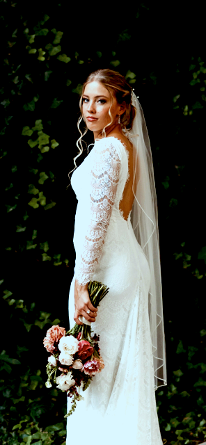 Bride in a white wedding dress holding a floral bouquet, standing against a dark green leafy background.
