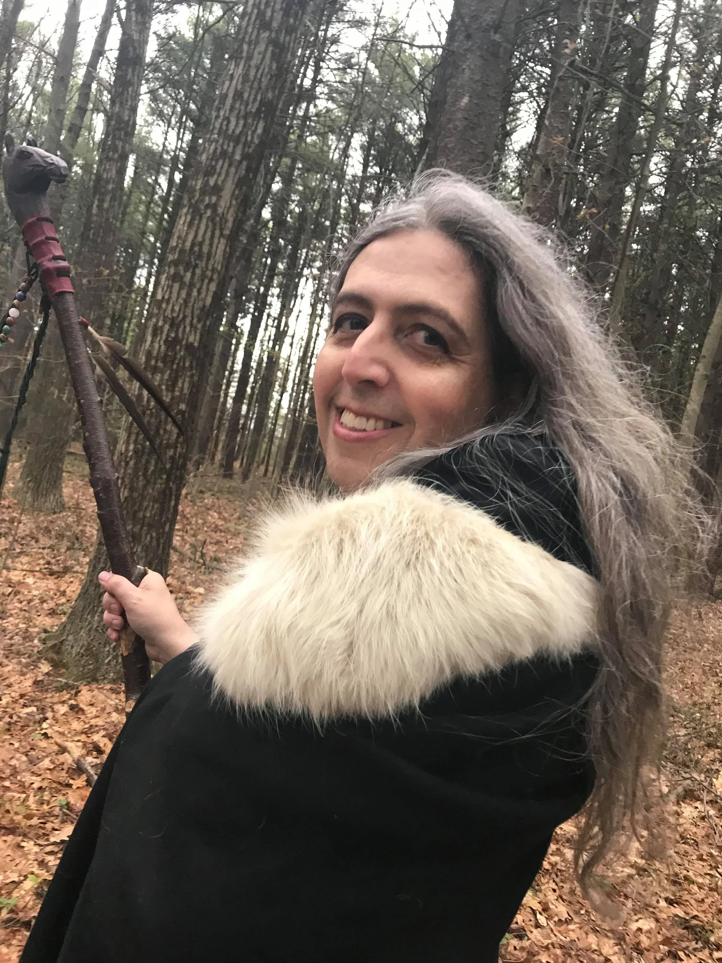 A woman with long gray hair smiling and holding a walking stick in a forest.
