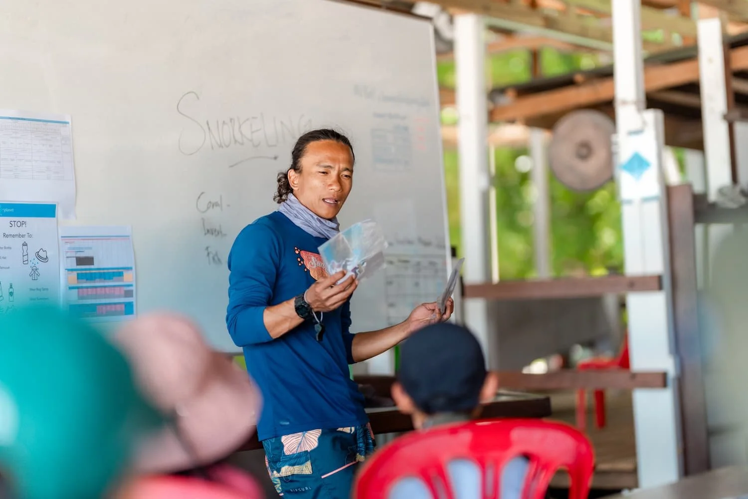 A man with long curly hair tied back, wearing a blue shirt and patterned shorts, stands in front of a whiteboard and speaks to children sitting in red chairs. The whiteboard has handwritten notes and laminated charts are posted nearby. The setting appears to be outdoors or in a semi-open structure with greenery visible outside.