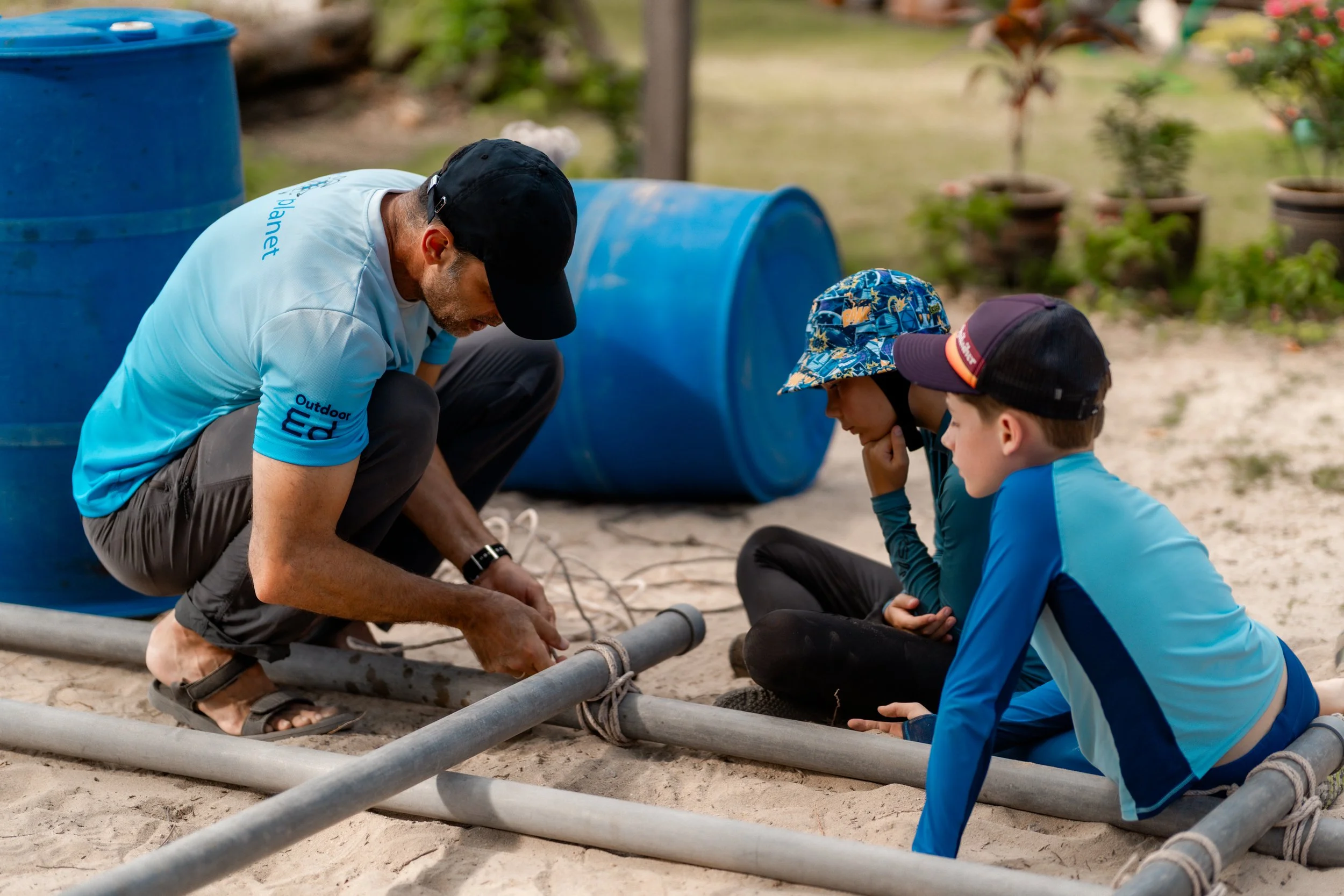 Man and two boys working on a project with pipes outdoors, with potted plants in the background.