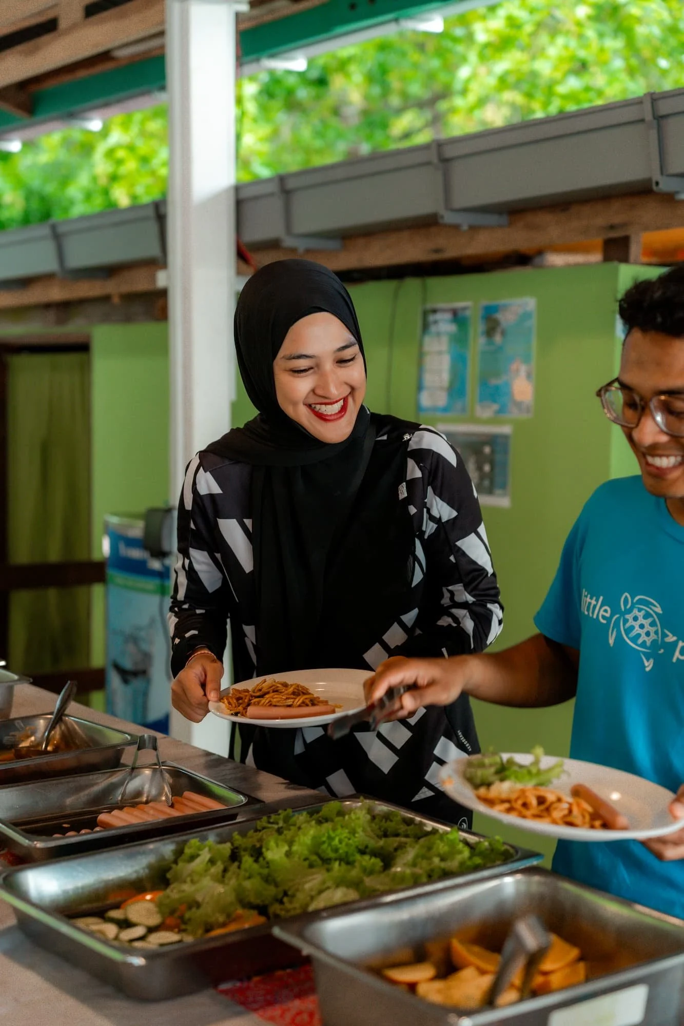 Two people serving food at a buffet, with trays of salad, pasta, and other dishes in front of them, and green and wooden background.