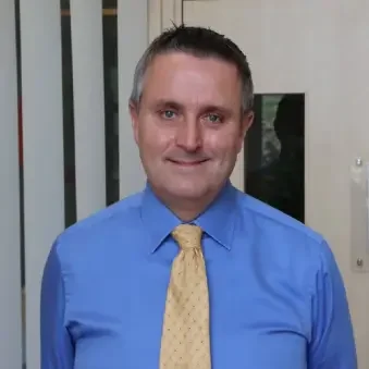 A man wearing a blue shirt and a yellow tie, standing indoors with vertical blinds and a window behind him.