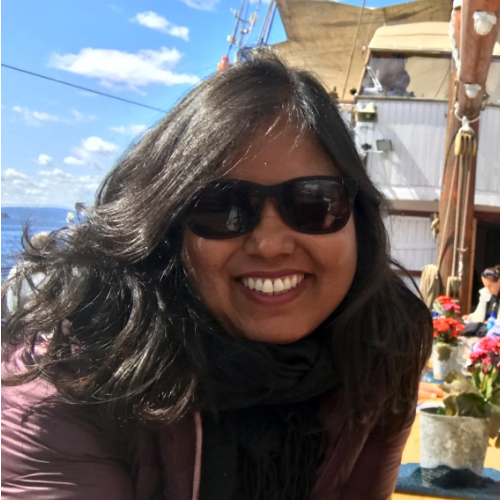 A woman with long dark hair wearing sunglasses smiling outdoors near a dock with boats and a building, under a partly cloudy sky.