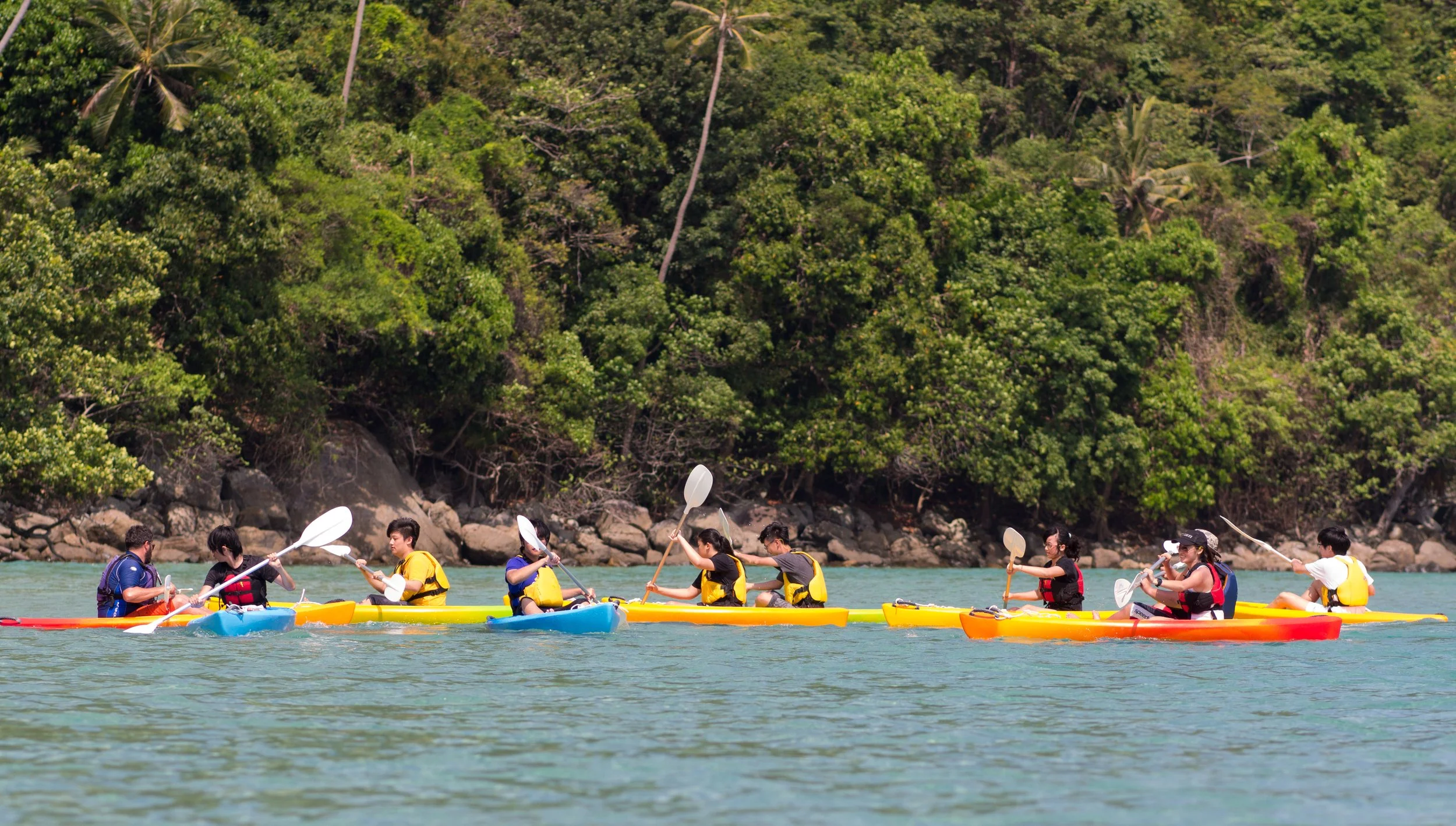A group of people kayaking on a river, surrounded by lush green trees and rocky shoreline.