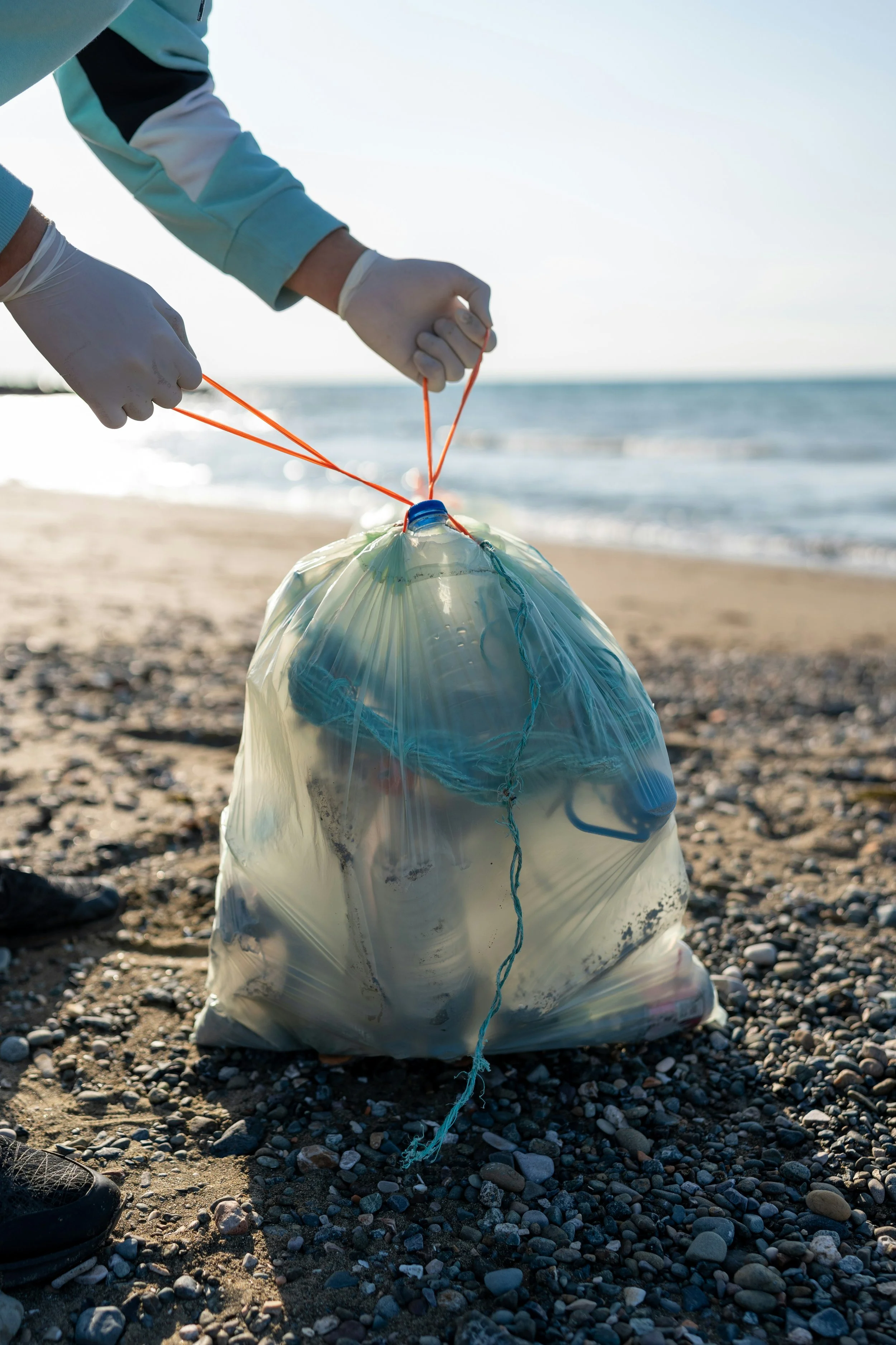 People collecting trash in a plastic bag on a pebble beach by the ocean, wearing gloves and using orange strings for gathering debris.
