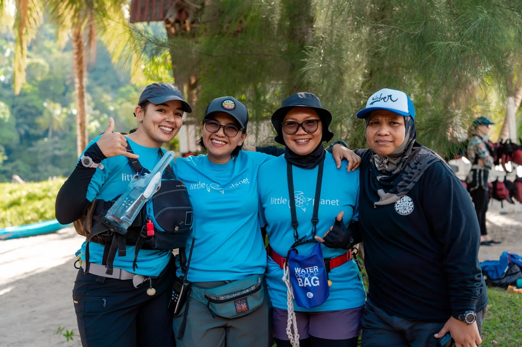 Four women standing together outdoors with green trees in the background, smiling and posing for a photo. They are wearing outdoor gear and blue shirts that say 'little planet,' suggesting they are part of a group or event.