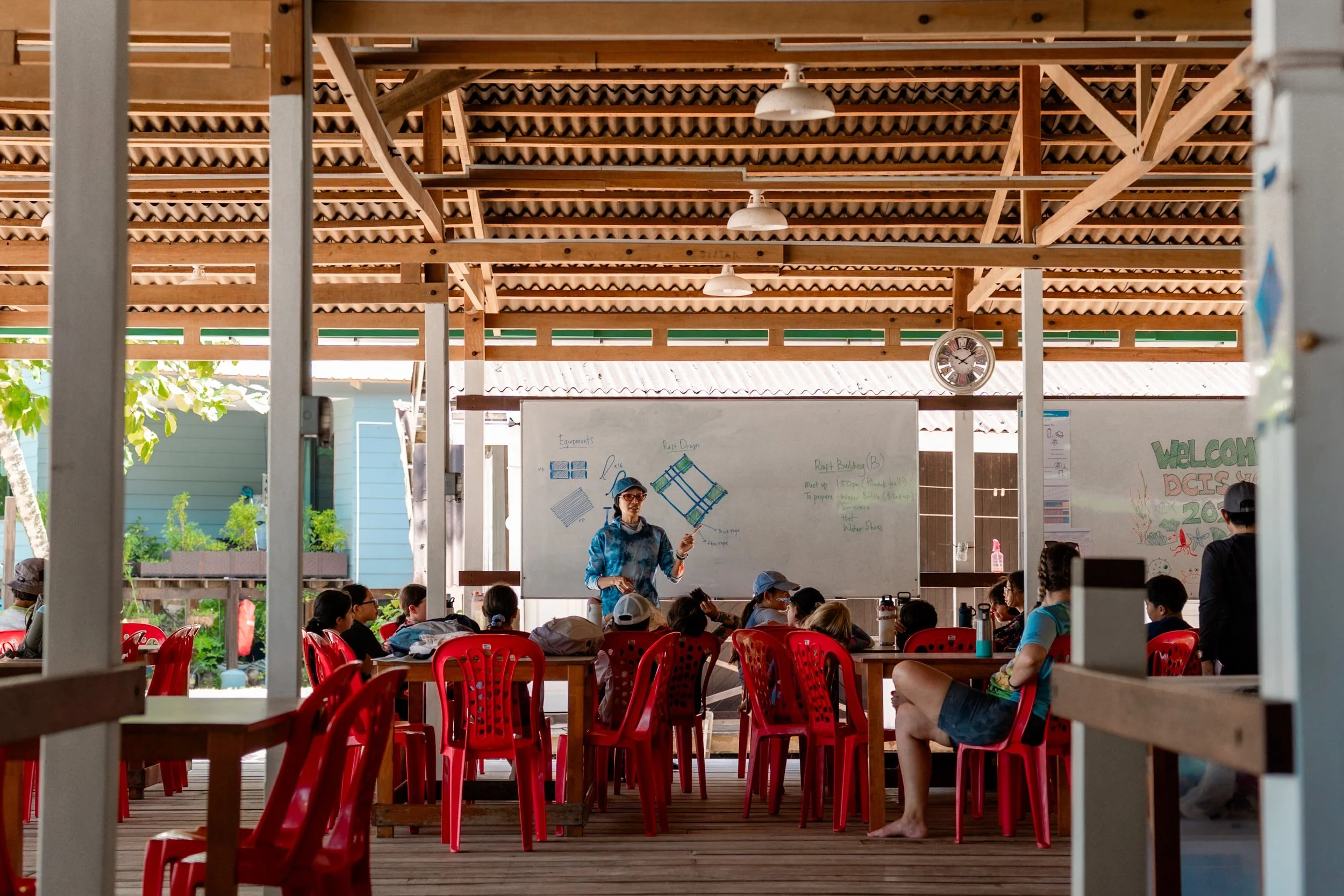 A group of children seated on red chairs inside a wooden pavilion, listening to a woman giving a presentation. The scene is viewed through an open window or doorway, with visible wooden structure and a whiteboard behind the speaker.