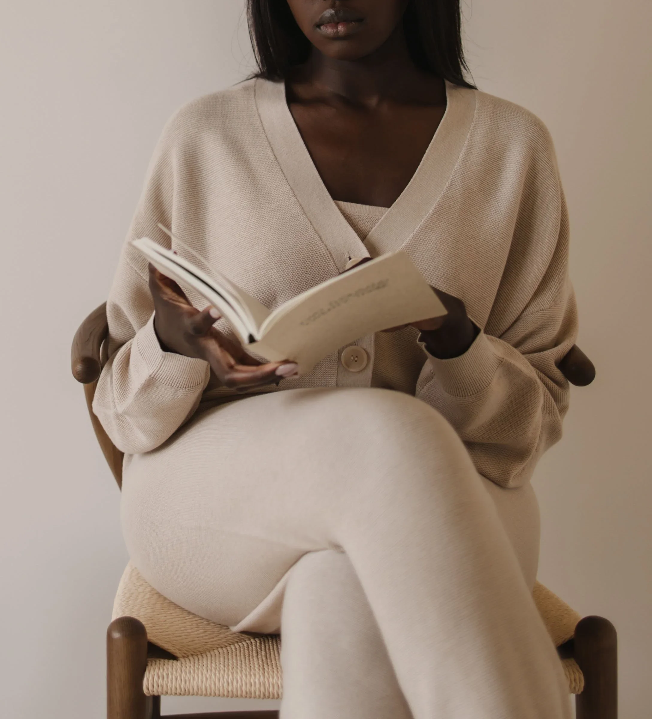 A woman sitting on a wooden chair, wearing a beige cardigan and holding an open book, with a plain light-colored wall in the background.