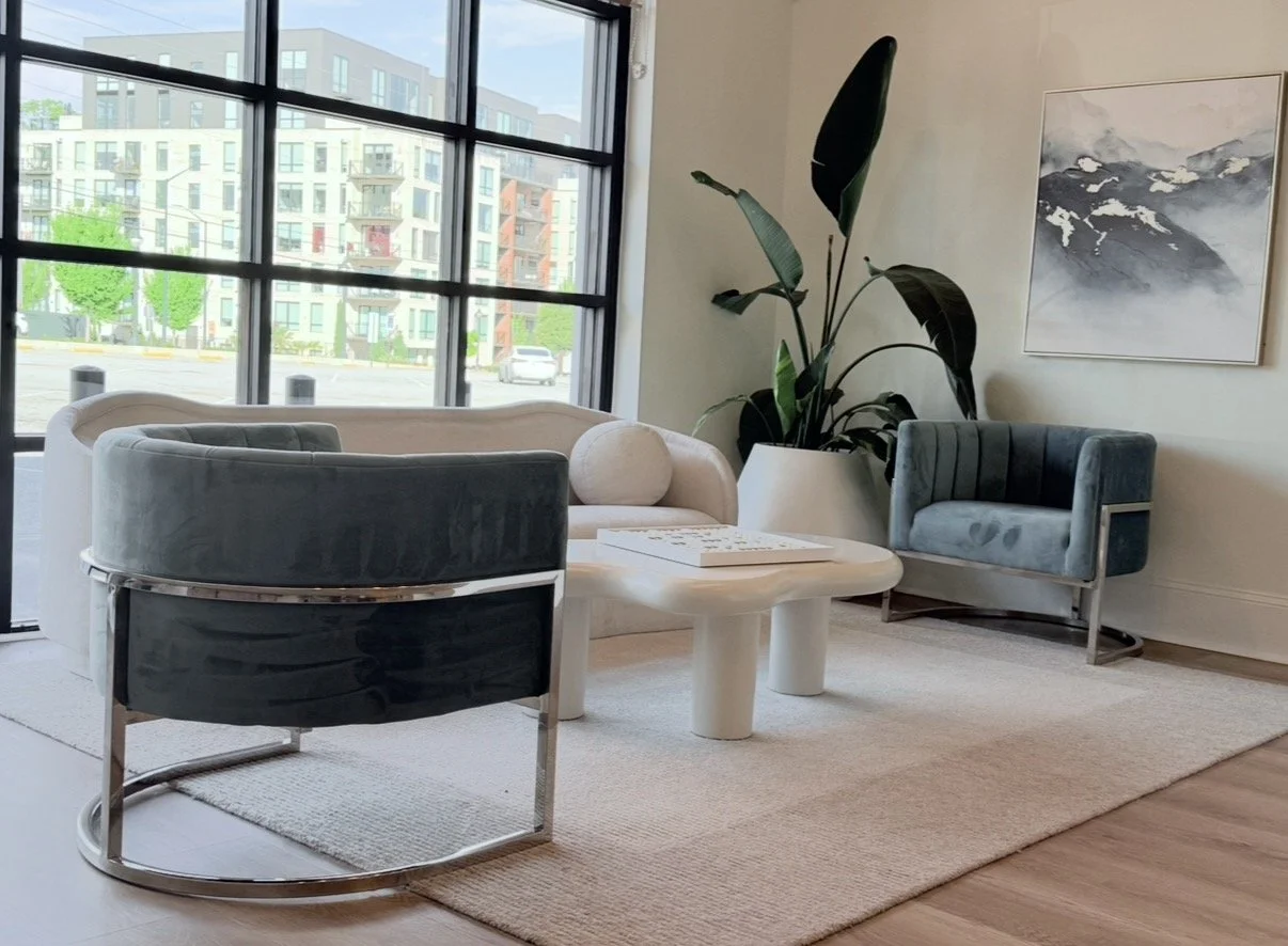 Modern living room with white sofa, two blue velvet armchairs, a white coffee table, a large houseplant, and a framed mountain landscape painting, with large windows showing a cityscape outside.