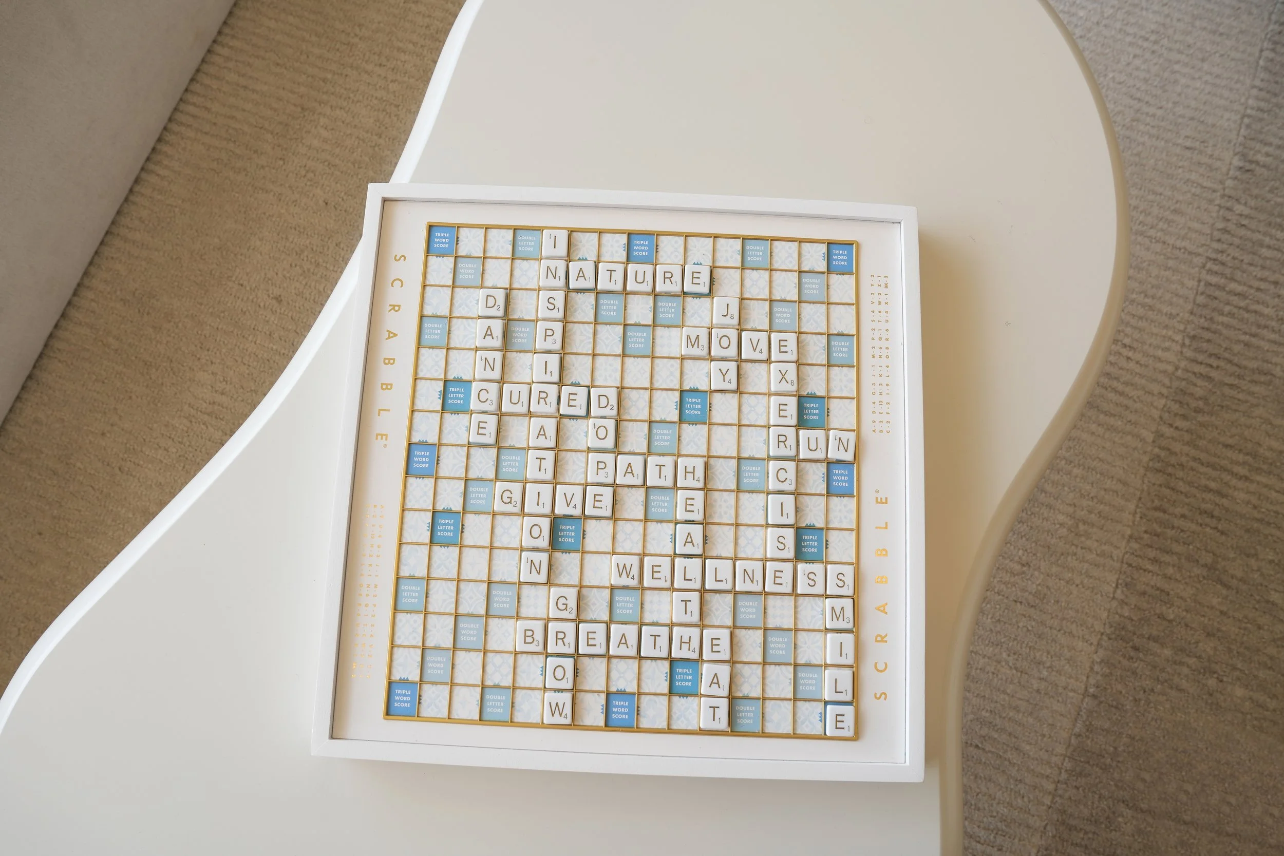 A Scrabble game board on a white table with words like 'NATURE', 'CURED', and 'BREATHE' spelled out with letter tiles, surrounded by a beige carpet.