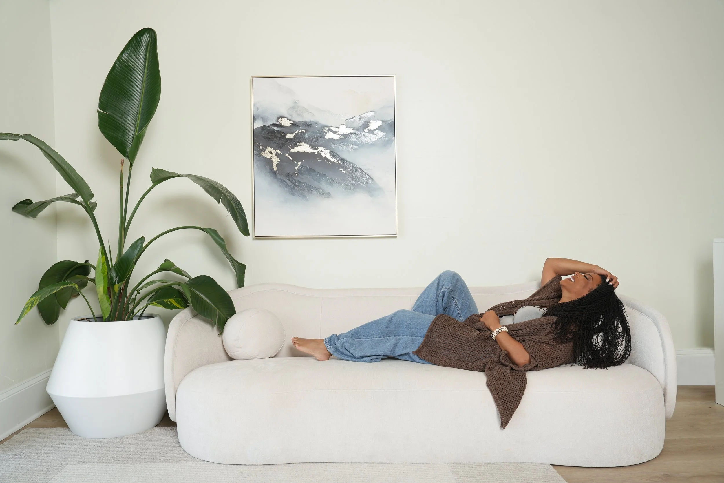 Woman with braided hair relaxing on a cream-colored sofa in a living room, with a large green houseplant in a white pot and a framed mountain painting on the wall behind her.