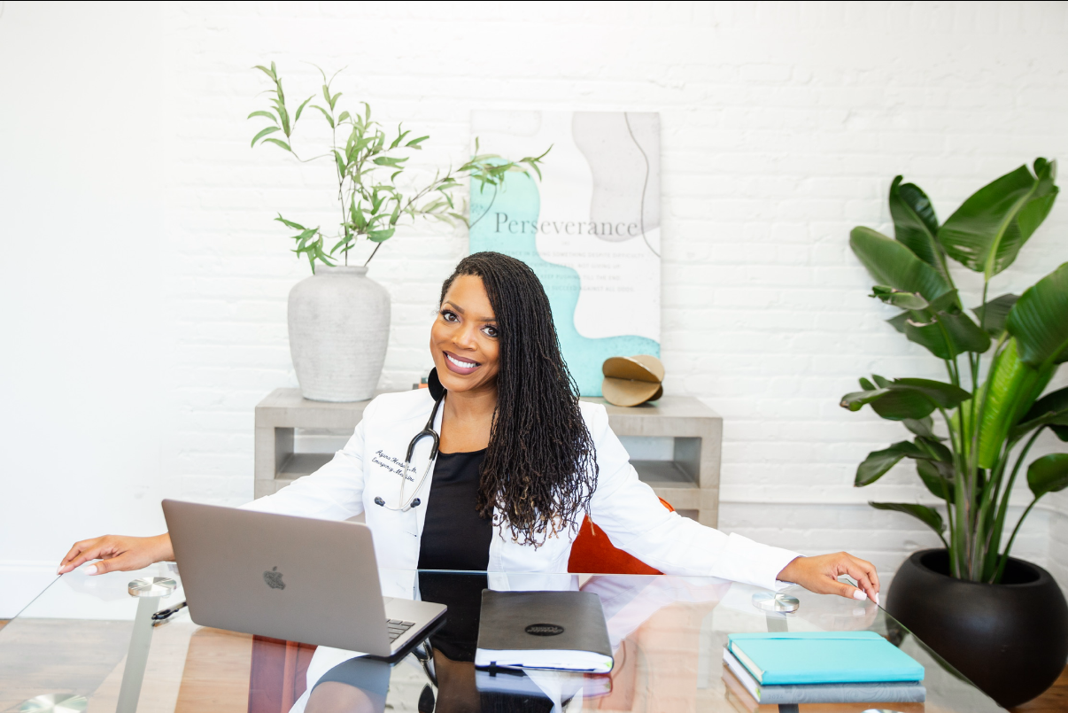 A female doctor smiling and sitting at a glass desk with a laptop, notebook, and books, in a bright modern office with green plants and artistic decor.