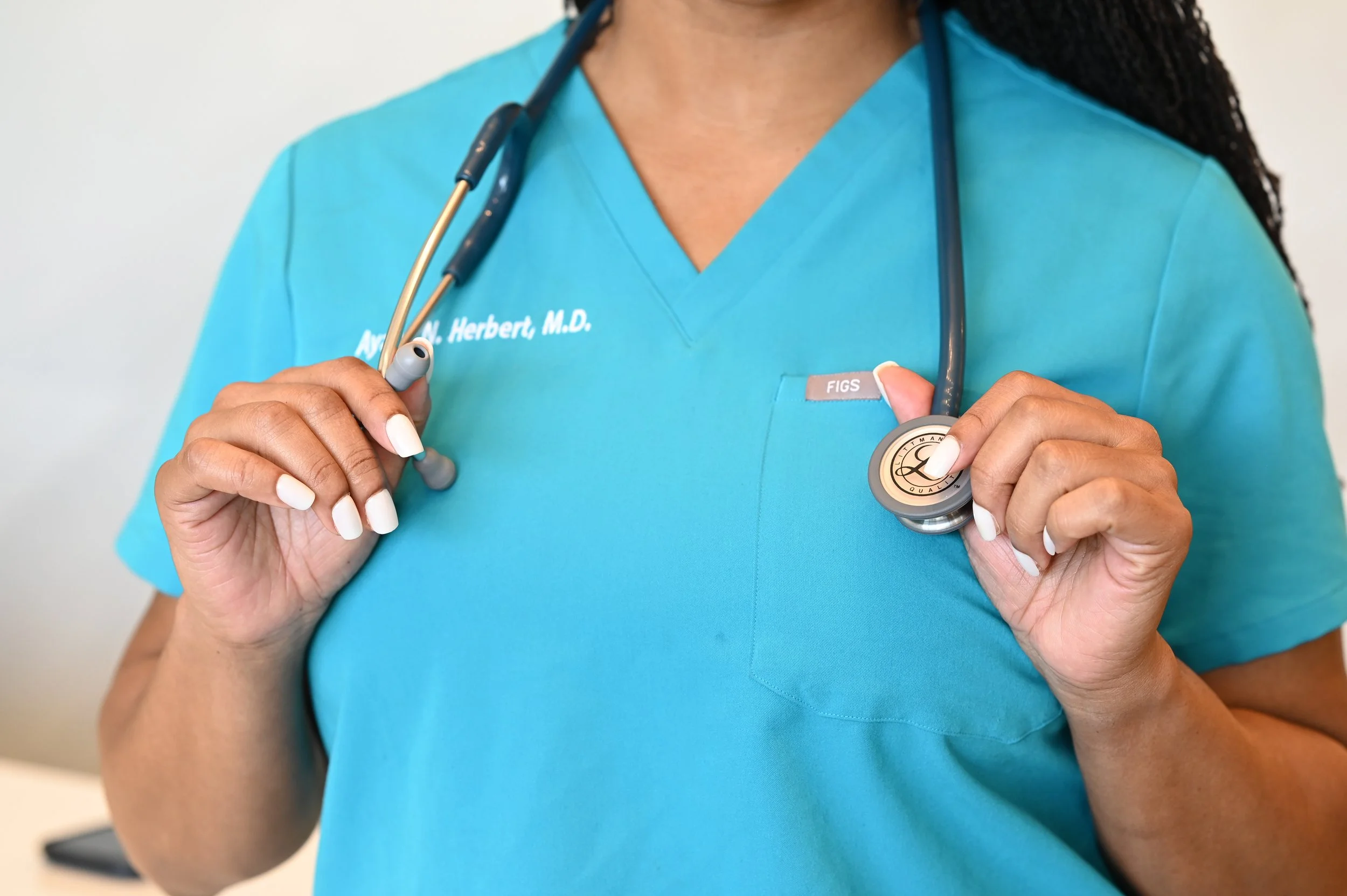 Close-up of a healthcare professional wearing a teal scrub top with a stethoscope around her neck. The professional is holding a badge or name tag in her right hand.