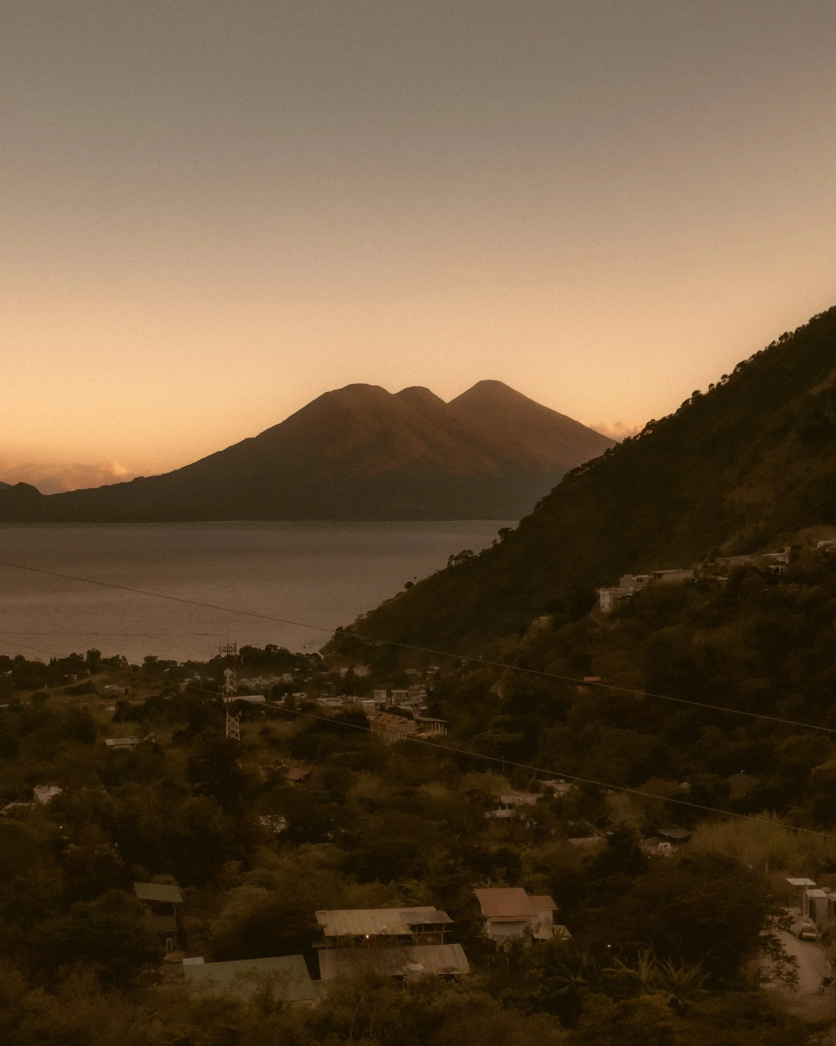 Sunset over a mountain range with a small town and houses at the foot of the hills, some trees in the foreground, and a calm body of water in the valley.
