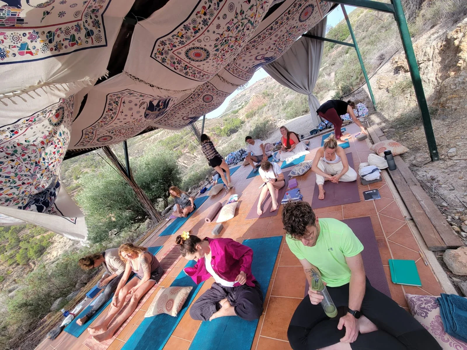 Group of people practicing yoga outdoors on a balcony with mountain views, colorful blankets and pillows, and yoga mats.