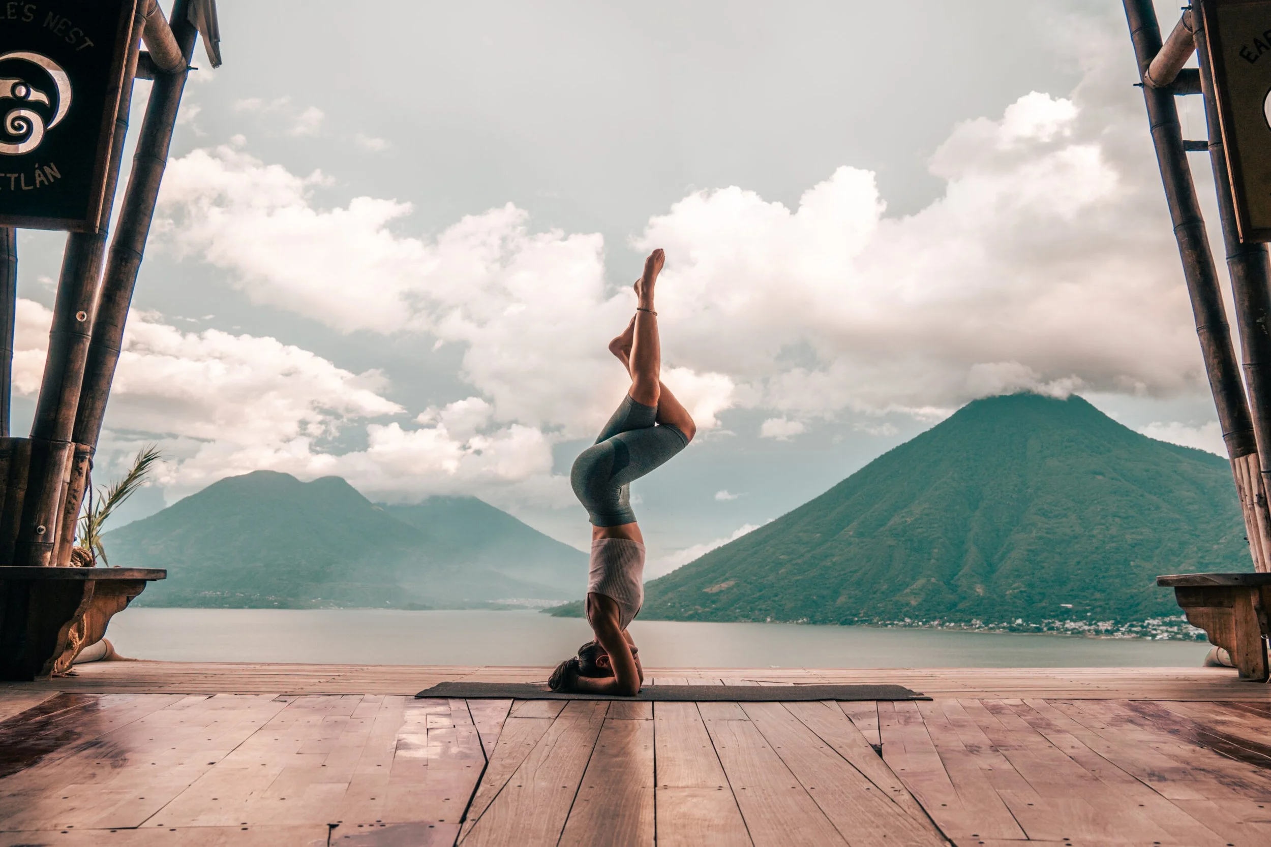 A person practicing yoga in a headstand pose on a wooden deck, with a scenic mountain and lake landscape in the background.